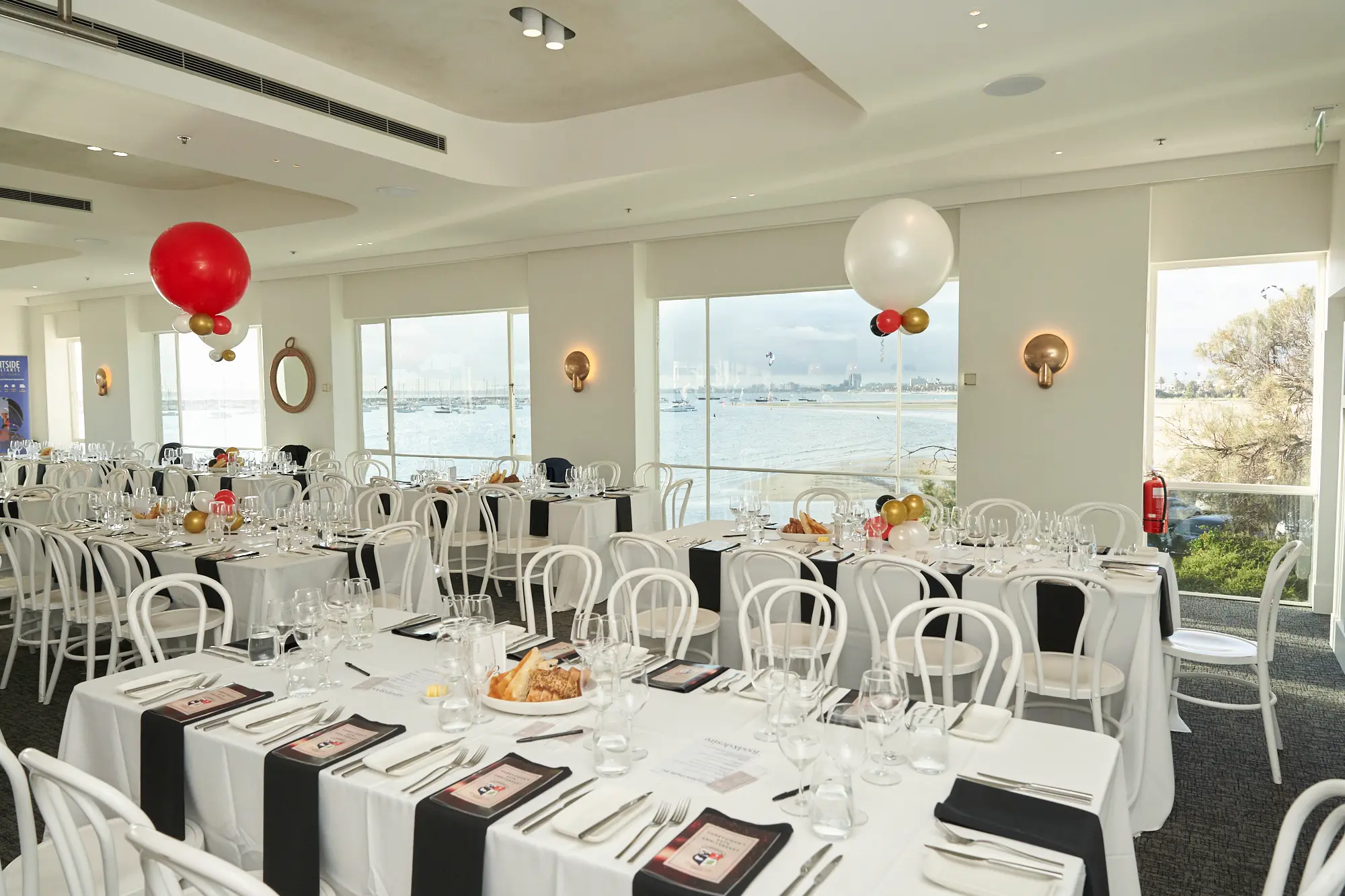garryowens 65th anniversary ball waterfront venue overview decor A bright waterfront event space is fully prepared for Garryowen’s 65th Anniversary Ball, featuring long dining tables arranged with white tablecloths, white chairs, black runners, polished glassware, and uniform place settings. Floor-to-ceiling windows showcase panoramic views of the calm bay with distant boats, flooding the room with natural light. Red and white balloon clusters add festive color to the modern interior with white walls and a curved ceiling. The meticulous decor, clean lines, and serene coastal backdrop create a sophisticated atmosphere of anticipation before guests arrive for the awards night celebration.
