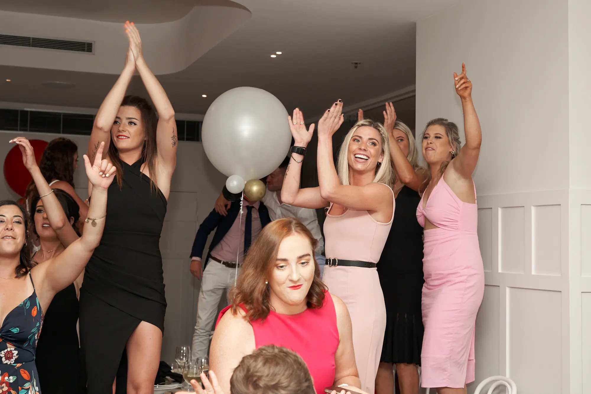 garryowens 65th anniversary ball women cheering dance floor A lively group of women enthusiastically cheer and raise their hands during Garryowen’s 65th Anniversary Ball. Dressed in elegant cocktail attire—black, dark pink, light pink, and red dresses—they celebrate on a polished hardwood floor amid formal dining tables with white chairs and tablecloths. A man in a blue suit and large decorative balloons add context to the festive indoor venue. The bright, warm lighting highlights their joyful expressions and energetic gestures, capturing a high-energy, electric moment of celebration, camaraderie, and pure enjoyment at this milestone awards night and anniversary gala.