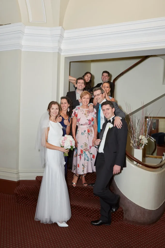 Masha and Marcus pose with family and guests on the grand staircase landing inside Melbourne’s historic Old Treasury Building. The bride stands on the lower left holding her pink-and-white bouquet, while the groom leans playfully forward. Seven other family members and guests are tiered behind them, all smiling at the camera. The elegant 19th-century staircase features a deep red patterned carpet, dark wooden railing, classic white cornices, wall panels, and a table with a floral arrangement. Soft natural light highlights the joyful, celebratory atmosphere, capturing a lively, fun, and timeless group moment in this heritage venue.