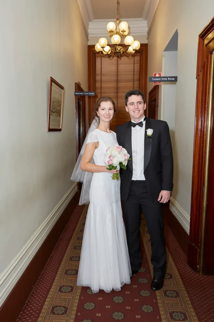 Masha and Marcus stand side-by-side in an elegant hallway of Melbourne’s historic Old Treasury Building, smiling joyfully at the camera. The bride wears a cap-sleeved lace gown and veil, holding a pink and white bouquet, while the groom stands in a classic black tuxedo with a white boutonnière. Behind them, dark wood wainscoting, a red-and-gold runner carpet, and signage for the Thomas Hyde Room and Margaret Craig Room highlight the venue’s heritage. Soft natural light and a brass chandelier create a warm, timeless atmosphere for this refined pre-ceremony portrait.