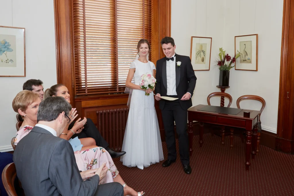 A quiet and intimate wedding ceremony at Melbourne’s Old Treasury Building, showing Masha and Marcus standing together as the groom reads their vows or official document. Masha holds a soft pink-and-white bouquet, while guests sit closely in the foreground, watching warmly and applauding. The room features dark wood wainscoting, botanical wall art, window blinds, and a traditional red carpet, creating an elegant registry office atmosphere. This heartfelt moment captures the couple’s refined style and the timeless charm of their Melbourne wedding.