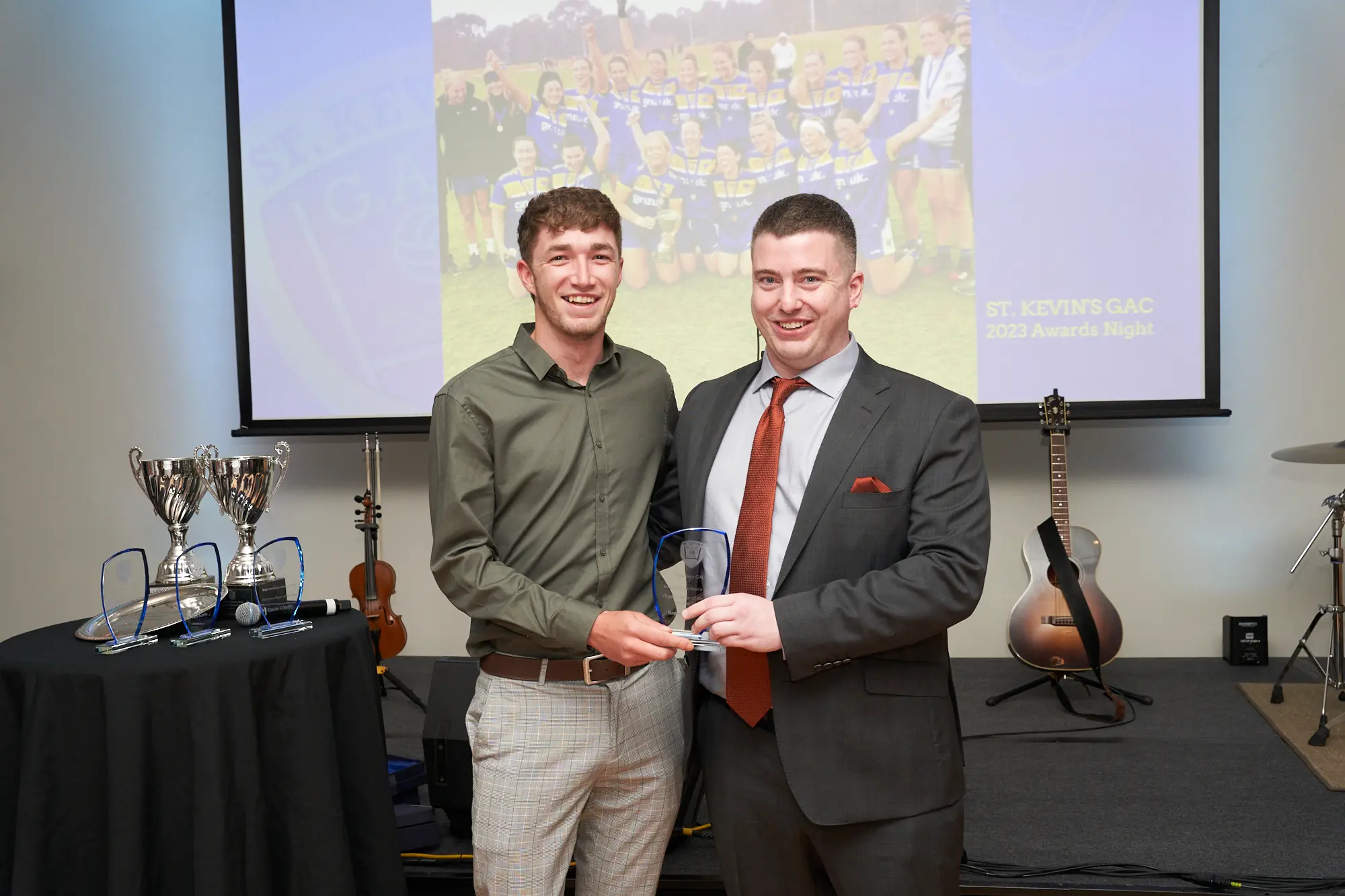 st kevins awards night award presentation on stage pullman melbourne albert park Two men smile as they pose on stage during St. Kevin’s Awards Night. The man on the left, wearing an olive-green shirt and grey checked trousers, holds a glass award plaque, while the man on the right, dressed in a dark grey suit and rust-orange tie, presents it. Behind them, a dark-covered table displays large silver trophies, and musical instruments including a guitar, cello, and drum set are visible. A large projection screen shows an image of a women’s sports team with the title “ST. KEVIN’S GAC 2023 Awards Night.” The atmosphere is formal, cheerful, and celebratory, reflecting a proud awards moment at Pullman Melbourne Albert Park.