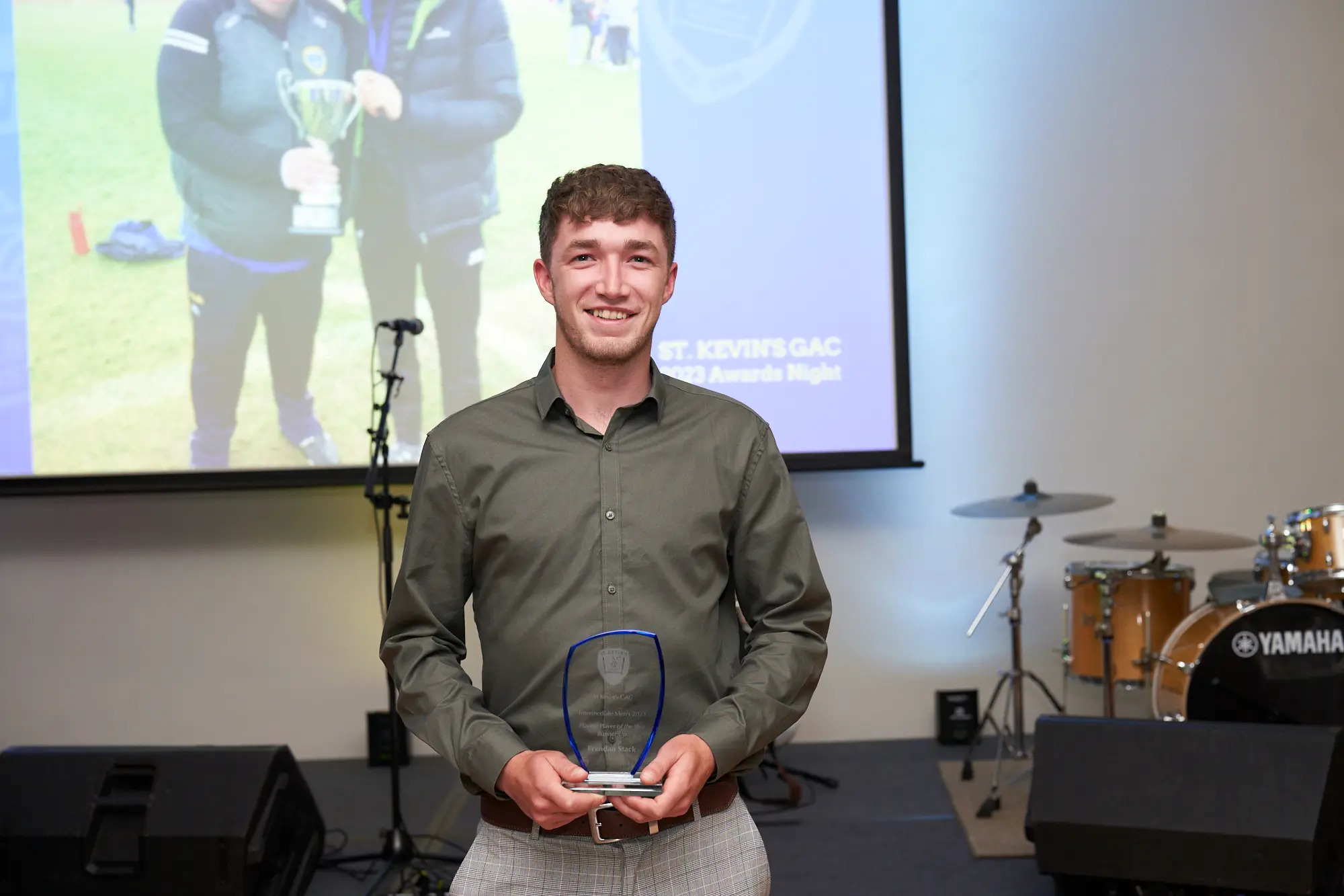 st kevins awards night award winner on stage pullman melbourne albert park A young man with short brown hair smiles proudly on stage during St. Kevin’s Awards Night. He is wearing an olive-green button-up shirt and grey patterned trousers while holding a clear, shield-shaped glass award plaque in both hands. Behind him, a large projection screen displays an image of two people holding a silver trophy, along with the text “ST. KEVIN’S GAC 2023 Awards Night.” To the right, a drum set with the Yamaha logo and other audio equipment are visible. The lighting focuses on the award recipient, creating a formal, celebratory, and uplifting atmosphere at Pullman Melbourne Albert Park.