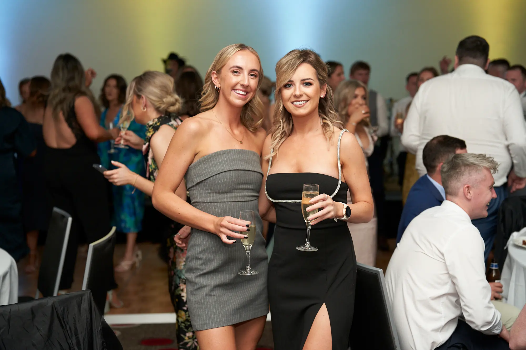 st kevins awards night capturing atmosphere guest moments women posing Two women pose closely together, smiling warmly at the camera during the annual St. Kevin's Awards Night. The blonde woman wears a grey strapless dress, while her friend wears a black dress with pearl-style shoulder straps, both holding champagne flutes. Around them, guests in formal attire are engaged in lively conversation, some standing, others seated at tables with white tablecloths. Warm ambient lighting combined with subtle blue stage lights creates a festive glow, capturing the joyful, social energy and celebratory atmosphere of the awards night, where friends and colleagues enjoy an elegant and memorable evening together.