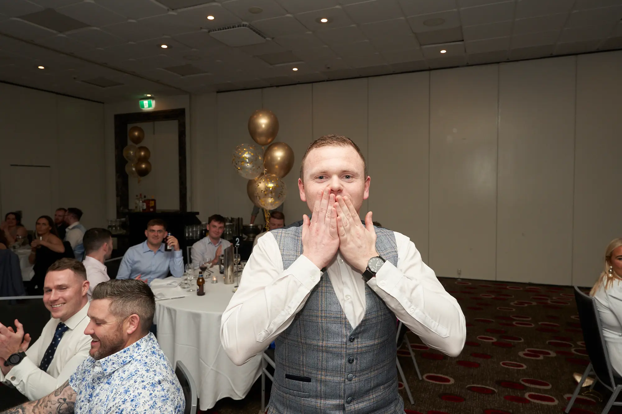 st kevins awards night guest reaction moment pullman melbourne albert park A young man in a white shirt and grey plaid waistcoat reacts with surprise during St. Kevin’s Awards Night at Pullman Melbourne Albert Park. Standing in the foreground, he covers his mouth with both hands, possibly in disbelief or excitement. Behind him, guests seated at round tables smile and applaud. The room features warm ambient lighting, white tablecloths, and festive gold and confetti-filled balloons near a dark archway. The mood is lively and celebratory, capturing a spontaneous and joyful reaction during the event’s highlight moment.