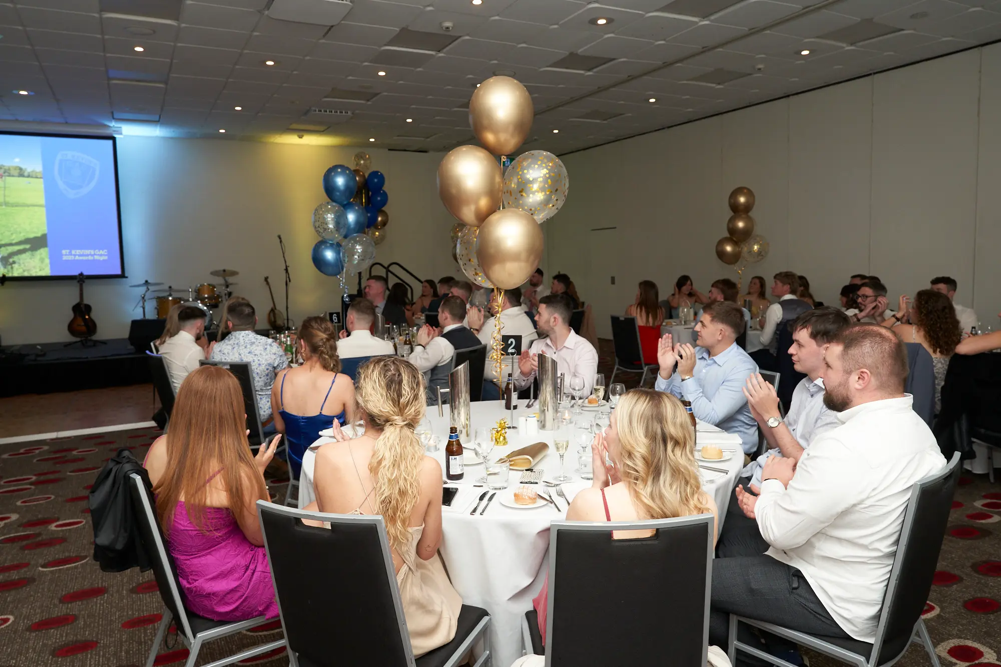 st kevins awards night guests applauding stage speech pullman melbourne albert park Guests seated at round tables applaud during St. Kevin’s Awards Night at Pullman Melbourne Albert Park. In the foreground, several people, including two men on the right, are clapping and looking toward the stage on the left. The banquet hall is filled with formally dressed attendees seated at tables set with white tablecloths, glassware, and tableware. The stage features a large screen displaying “ST. KEVINS” and musical instruments, including a guitar and drum set. Clusters of gold and blue balloons with confetti decorate the space, creating a formal, festive, and celebratory atmosphere.