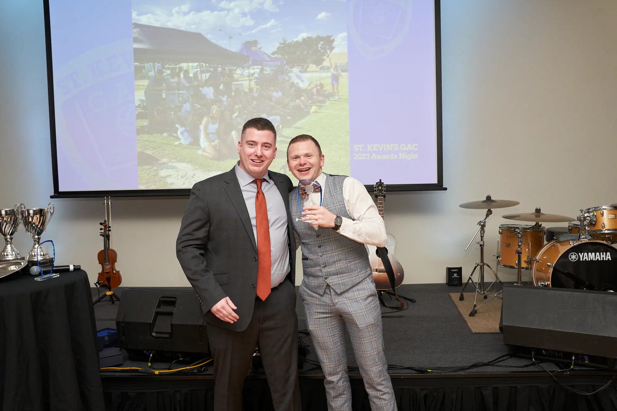 st kevins awards night pullman melbourne albert park award presentation stage photo two men posing A wide-shot photo capturing two men on stage during St. Kevin's Awards Night at Pullman Melbourne Albert Park. The man on the left wears a dark grey suit with a rust-orange tie, while the award winner on the right wears a grey checkered three-piece suit with a bowtie, holding a clear glass award plaque. Behind them, a large screen displays “ST. KEVIN'S GAC 2023 Awards Night,” and several trophies and musical instruments, including a guitar and drum kit, are visible on stage. The atmosphere is formal and celebratory, marking a proud moment of recognition.