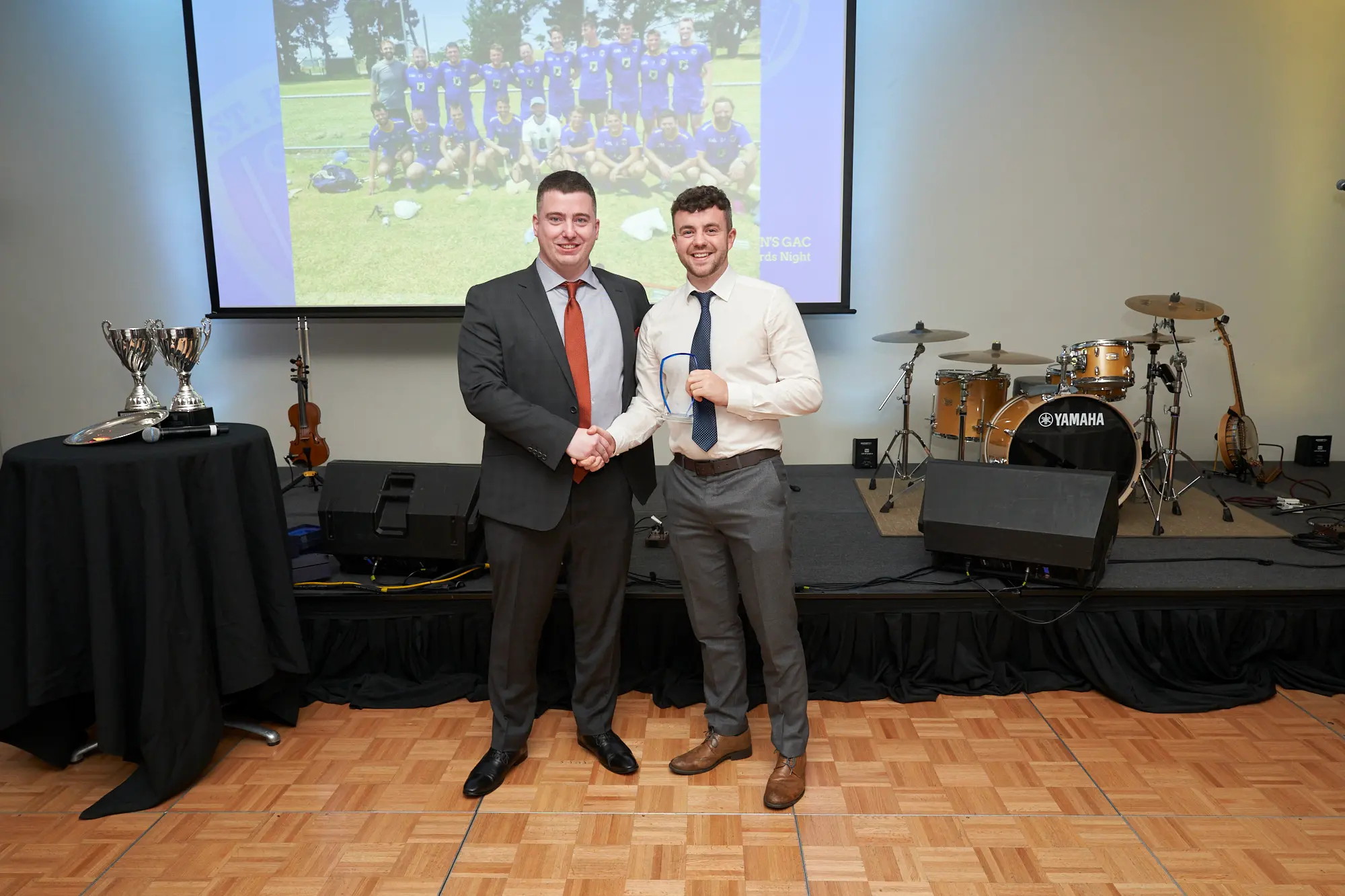 st kevins awards night pullman melbourne albert park award presentation winner posing with presenter on stage A wide-shot image of two men posing on stage during St. Kevin's Awards Night at Pullman Melbourne Albert Park. The man on the left, dressed in a grey suit with a white shirt and rust-orange tie, stands beside the award winner, who wears a light shirt, dark blue tie, and grey trousers while holding a clear, shield-shaped glass award plaque. Behind them, a projector screen displays a sports team photo with the text “ST. KEVIN'S GAC 2023 Awards Night.” A Yamaha drum kit, an acoustic guitar, and large silver trophies are visible on stage, contributing to the formal and celebratory atmosphere.