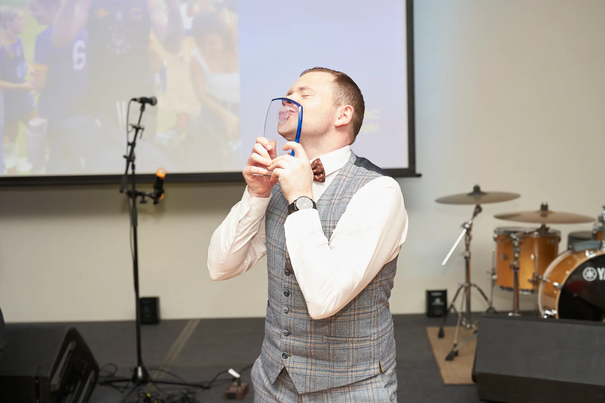 st kevins awards night pullman melbourne albert park award winner kissing trophy on stage A wide-shot image of an award winner celebrating on stage during St. Kevin's Awards Night at Pullman Melbourne Albert Park. The man, dressed in a grey plaid three-piece suit with a white shirt and patterned bowtie, is holding a clear, shield-shaped glass award plaque with blue accents and kissing it proudly. Behind him, a large screen displays an image from an outdoor event with the number “6” visible, along with a drum kit, speakers, and microphone stands. The atmosphere is joyful and emotional, capturing a heartfelt moment of triumph and celebration.