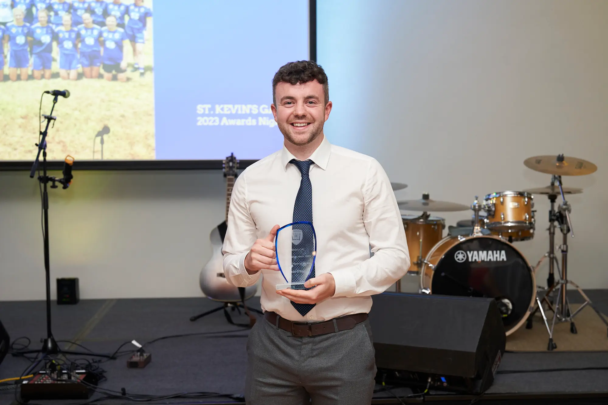 st kevins awards night pullman melbourne albert park award winner posing on stage with glass plaque A wide-shot image of a young man posing proudly on stage during St. Kevin's Awards Night at Pullman Melbourne Albert Park. He has short curly hair and is dressed in a white shirt, dark tie with blue accents, and grey trousers, smiling warmly while holding a clear, shield-shaped glass award plaque in both hands. Behind him, a projection screen displays a sports team photo with the text “ST. KEVIN'S GAC 2023 Awards Night.” A Yamaha drum set, an acoustic guitar, speakers, and audio equipment are visible on stage, enhancing the formal and celebratory setting.