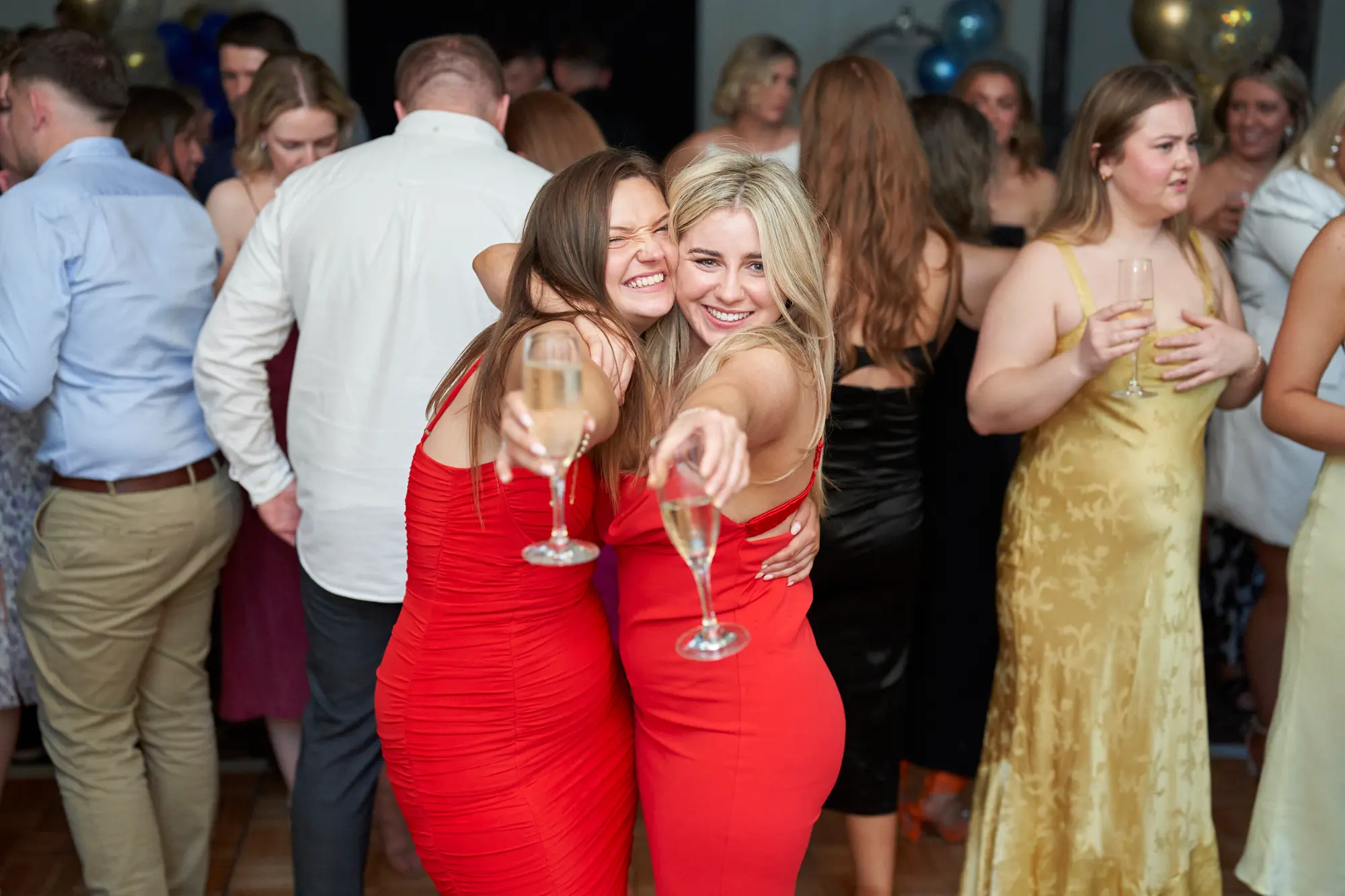 st kevins awards night pullman melbourne albert park candid celebration two women red dresses Two women in matching vibrant red dresses share a joyful hug and raise their champagne flutes toward the camera during St. Kevin’s Awards Night at Pullman Melbourne Albert Park. Their wide smiles and laughter capture a moment of pure celebration on the wooden dance floor, surrounded by mingling guests in formal attire. Warm lighting and festive details like gold balloons and sparkling drinks enhance the lively, elegant energy of the evening, evoking the spirit of camaraderie and joy that defines this annual awards celebration.