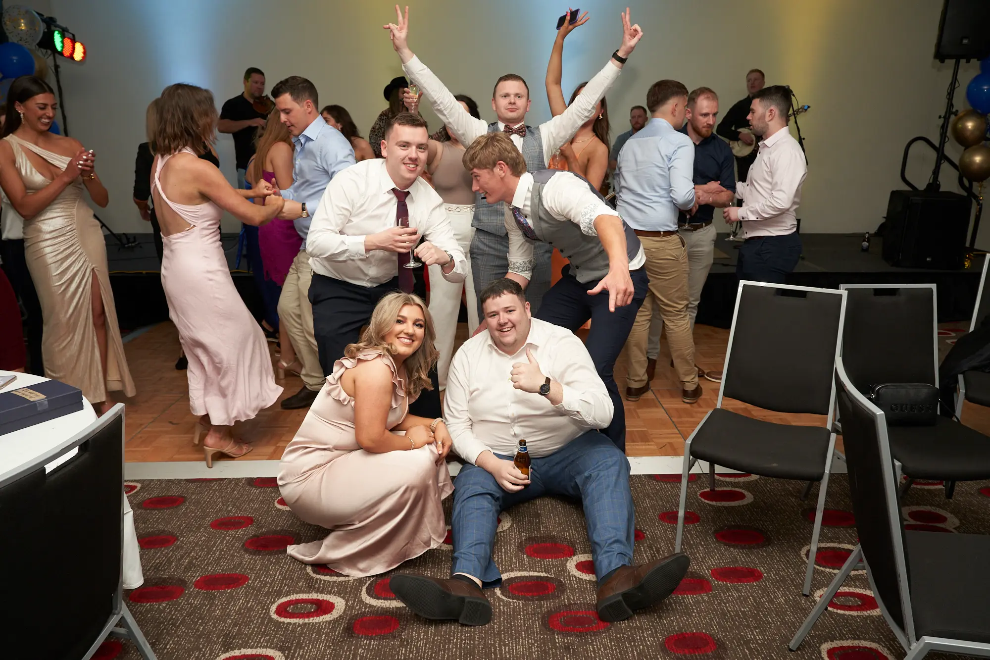 st kevins awards night pullman melbourne albert park candid dance floor celebration A candid wide shot captures guests celebrating at St. Kevin's Awards Night. In the foreground, a woman in a beige satin dress kneels with a wine glass, while a man in a white shirt and jeans gives a thumbs-up with a beer. On the dance floor, attendees mingle, dance, and raise arms in joy. Gold, blue, and confetti balloons decorate the room, with a stage and musical instruments in the background. The lively expressions, laughter, and celebratory gestures convey a festive, social, and joyous post-awards party atmosphere.