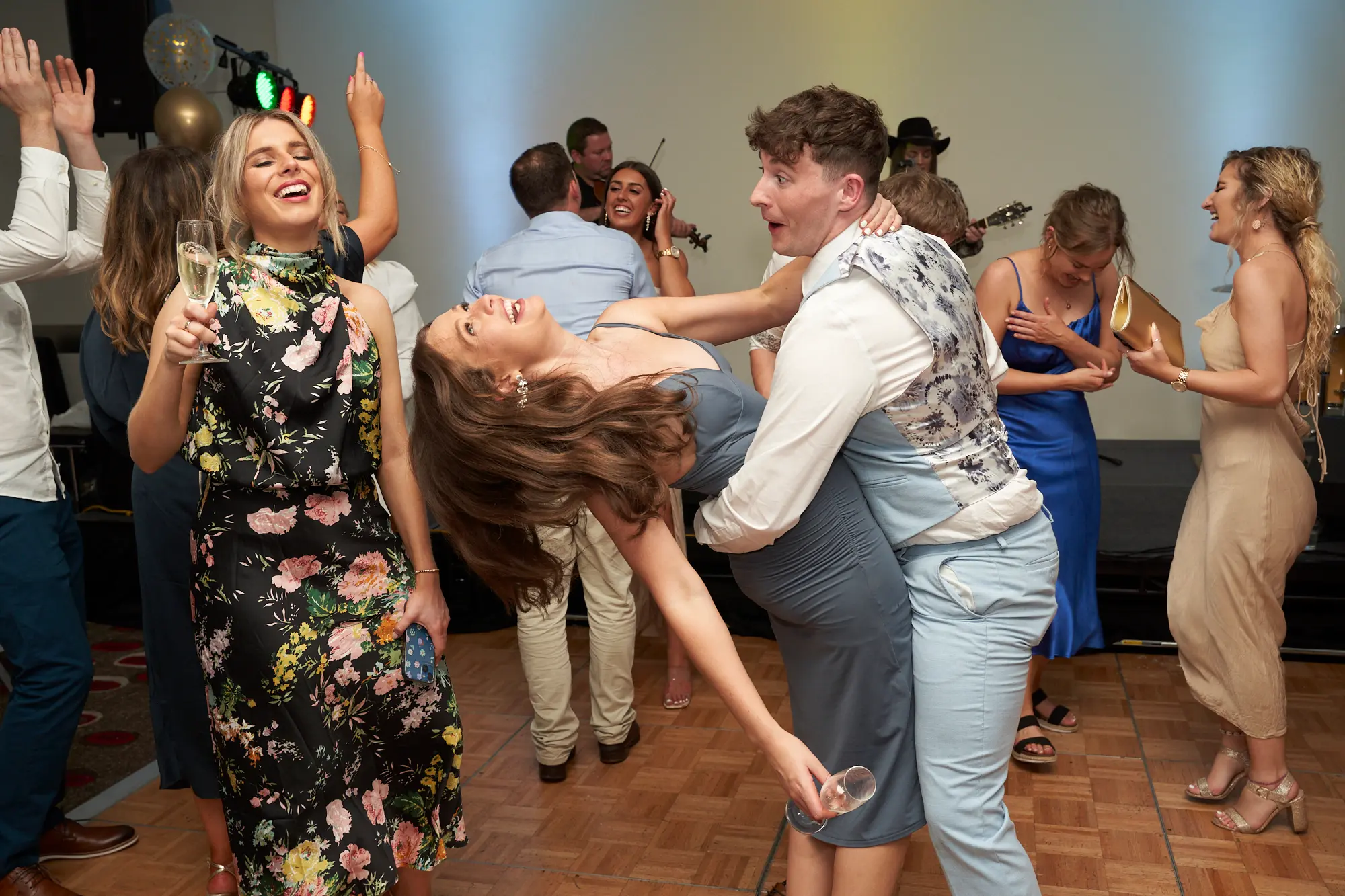 st kevins awards night pullman melbourne albert park candid dance floor energy A lively candid moment on the dance floor at St. Kevin's Awards Night. A man in a white shirt dips a woman in a grey dress mid-laugh, while others dance, cheer, and smile around them. Musicians play violin and guitar in the background, and balloons and colored lights decorate the ceiling. The image captures genuine joy, playful energy, and celebratory interactions among guests, reflecting the fun and spirited atmosphere of the awards night celebration.