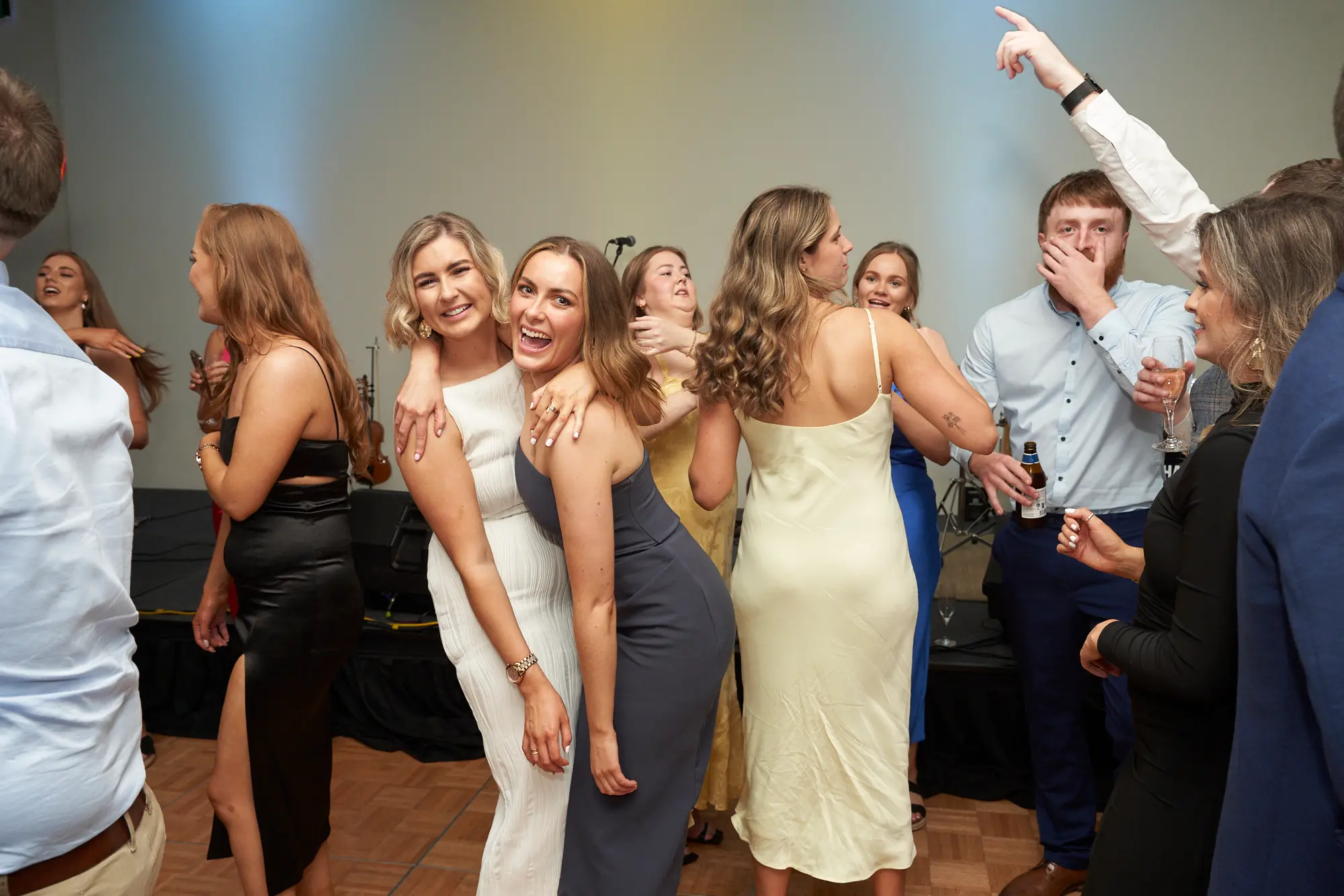 st kevins awards night pullman melbourne albert park candid dance floor laughter A candid wide shot captures guests enjoying the dance floor at St. Kevin's Awards Night. Two women in white and dark grey dresses pose closely, smiling with arms around each other. Nearby, a man in a light blue shirt laughs, pointing upward while holding a beer, and a woman in black joins in the fun. Other guests mingle in elegant attire. Wooden floors, a stage with musical instruments, and warm lighting set the scene. The vibrant expressions, laughter, and dynamic poses convey a joyful, lively, and celebratory post-awards party atmosphere.