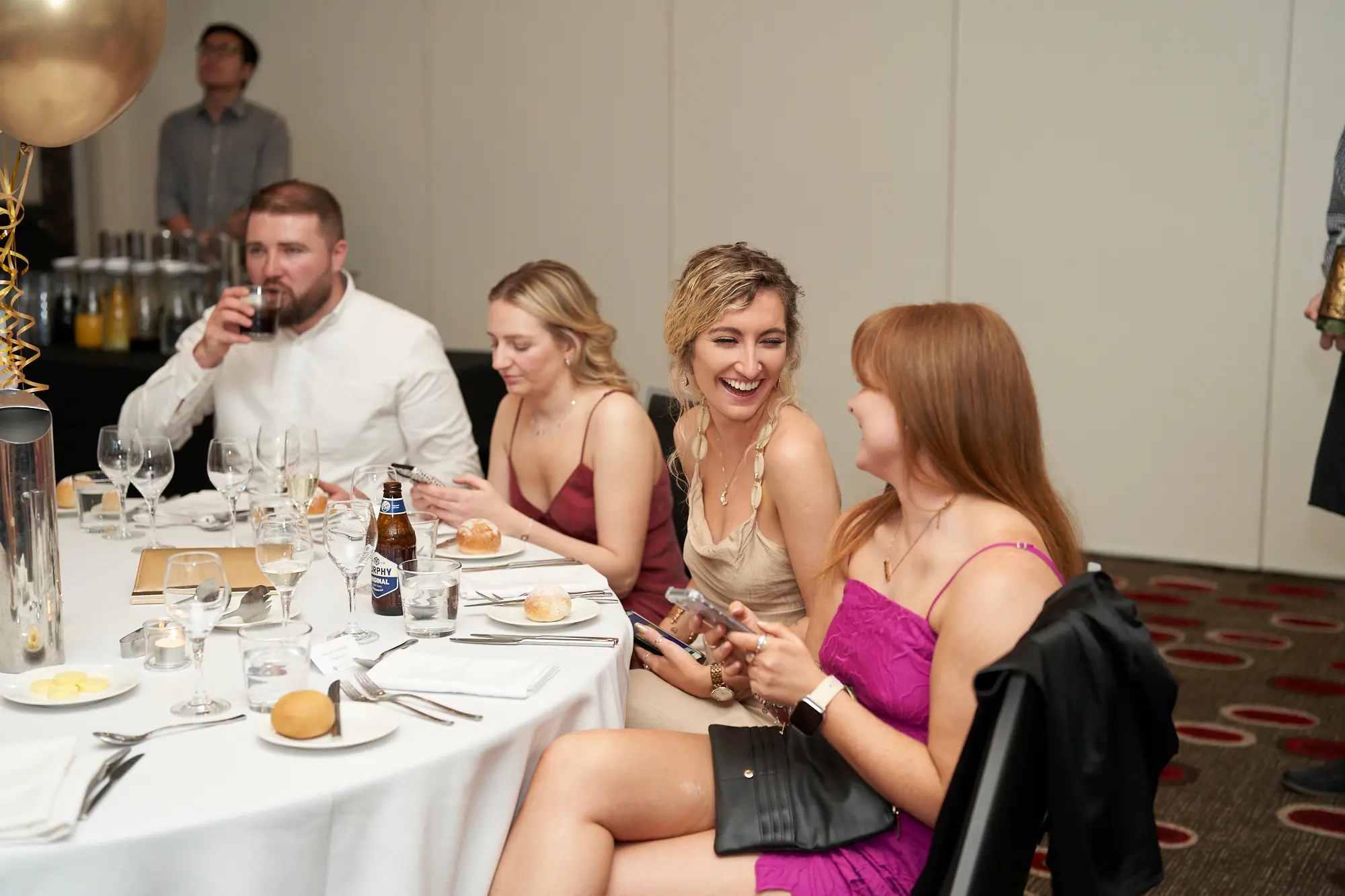 st kevins awards night pullman melbourne albert park candid dinner laughter A candid moment captures guests enjoying lively conversation and laughter at St. Kevin's Awards Night. Four individuals are seated at a round table: a bearded man sipping a drink, a woman in a maroon slip dress smiling at her phone, a woman in beige laughing with her head tilted back, and a red-haired woman in a bright fuchsia dress smiling at her companion. The white-tablecloth-covered table features glassware and bread rolls, with a gold balloon overhead. The image conveys warmth, friendship, and the joyful, relaxed atmosphere of a celebratory awards night dinner.