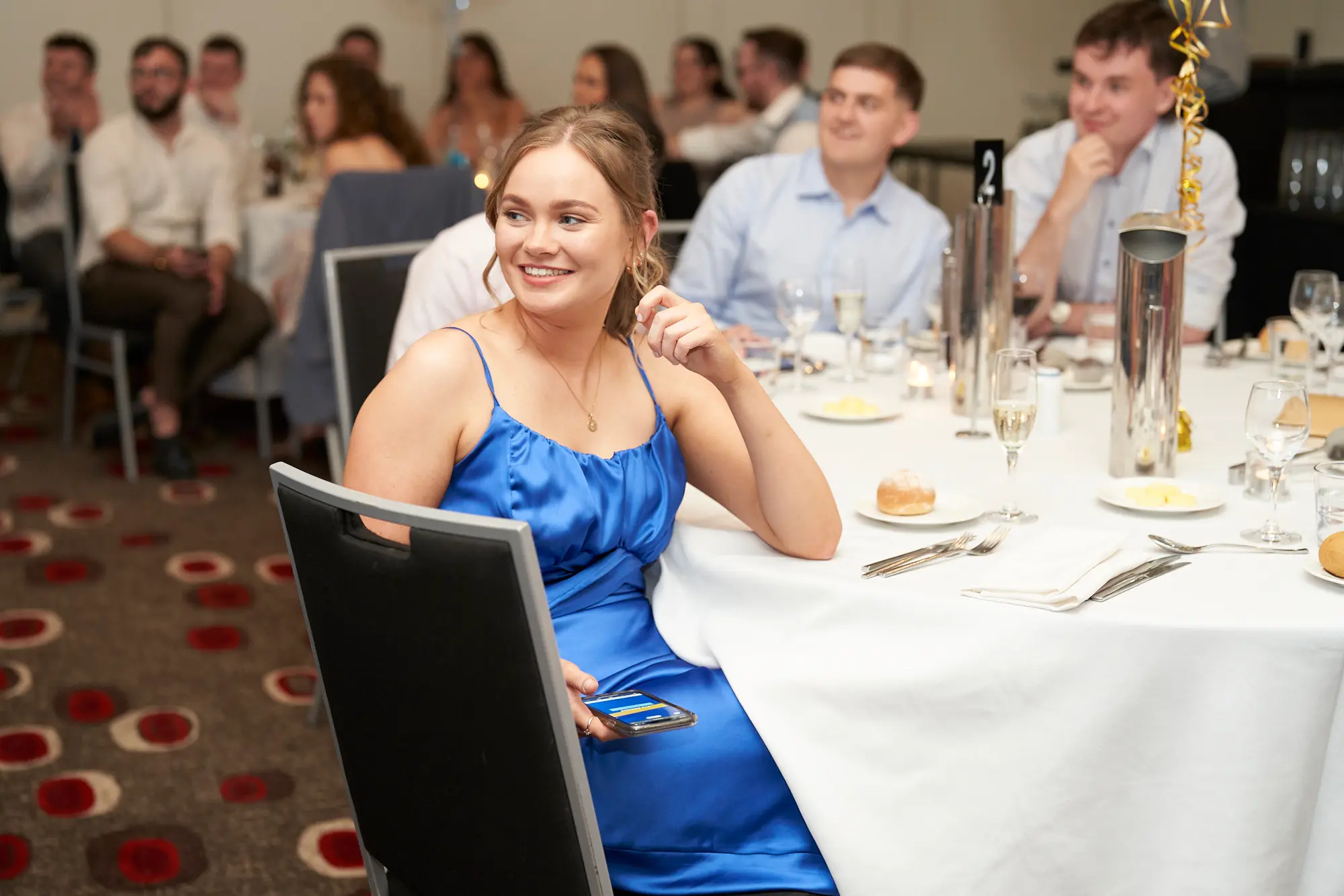 st kevins awards night pullman melbourne albert park candid guest engagement A candid moment captures a young woman in a royal blue satin dress seated at a round table during St. Kevin's Awards Night. She smiles broadly, resting her chin on her hand, engaged by an off-camera moment. Around her, guests at nearby tables chat and observe the event, with drinks and metallic table centerpieces visible. Warm ambient lighting and gold balloon decorations create a festive atmosphere. The image conveys genuine joy, attentiveness, and the celebratory spirit of a formal awards night, highlighting candid interactions and relaxed engagement among attendees.
