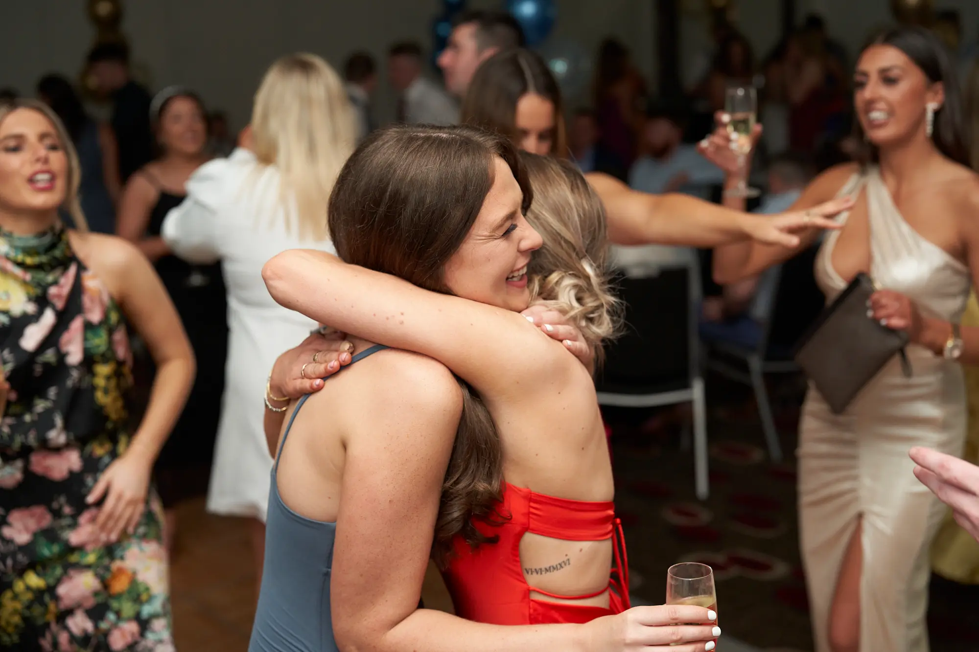 st kevins awards night pullman melbourne albert park candid hug celebration A candid photo captures two women sharing a heartfelt hug during St. Kevin's Awards Night. One wears a blue-grey slip dress with her back to the camera, while the other in a vibrant red dress laughs with eyes closed. Guests in the background, dressed formally, hold drinks and socialize amid gold and blue balloon decorations. The warm lighting, lively interactions, and genuine laughter convey a joyful, celebratory atmosphere, highlighting the emotional and festive energy of the post-awards party.