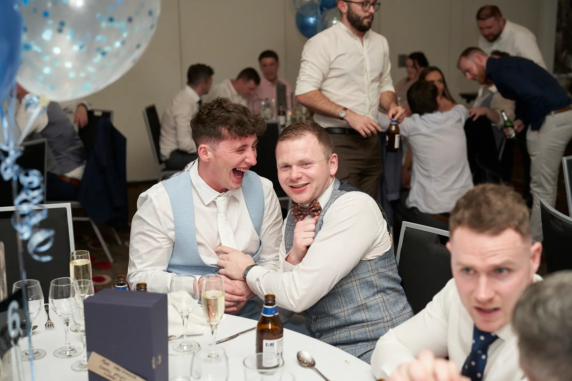 st kevins awards night pullman melbourne albert park candid laughter fest A candid photo captures a lively moment at St. Kevin's Awards Night, with two men at a dinner table sharing a hearty laugh. One in a white shirt and light blue waistcoat leans back, mouth open mid-laugh, hand resting on his friend’s shoulder, while the other in a grey checkered waistcoat and maroon bowtie smiles broadly at the camera. The table is set with plates, glassware, cutlery, and beer bottles, with festive blue and confetti balloons nearby. The image conveys joy, camaraderie, and the playful energy of a celebratory awards night dinner.
