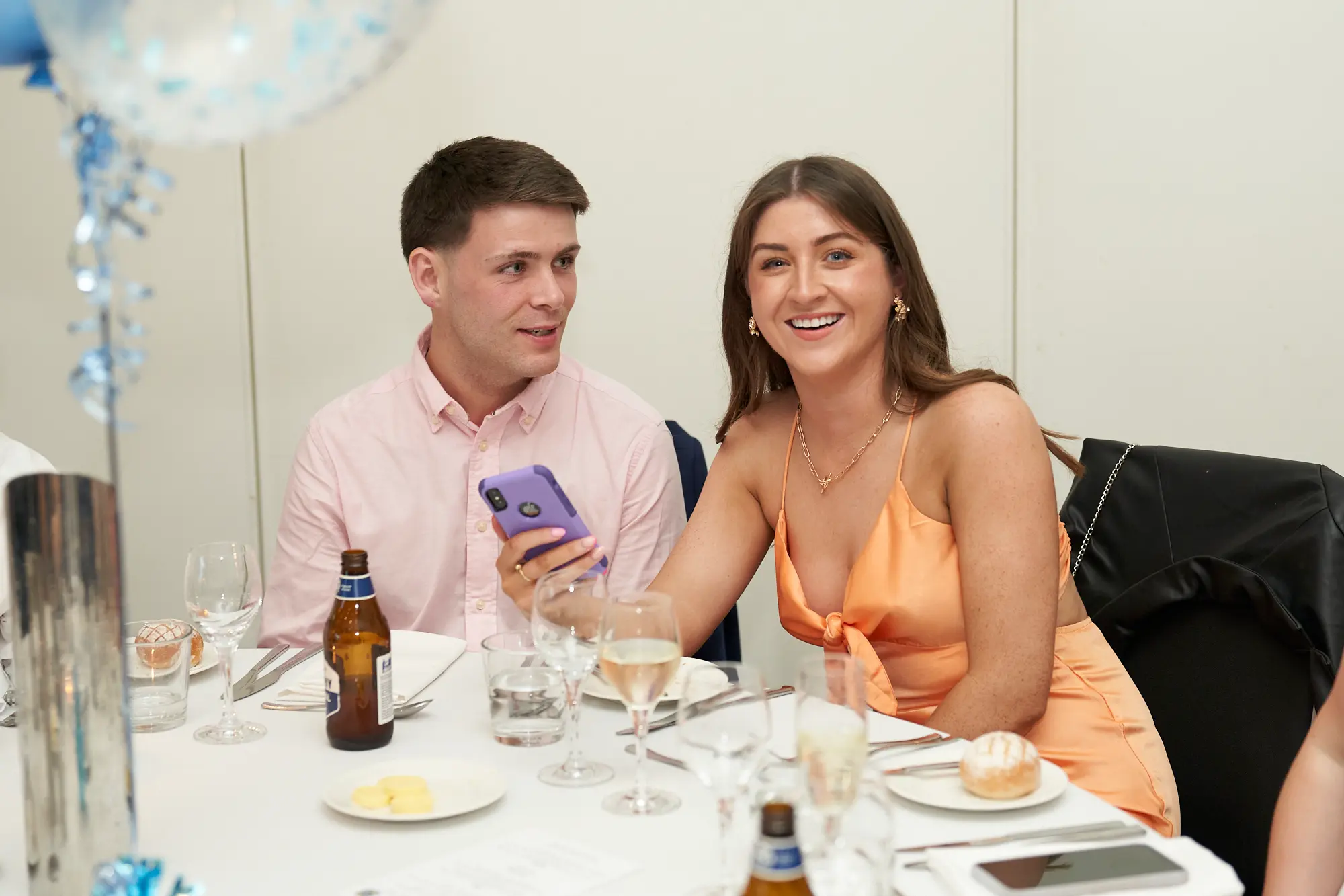 st kevins awards night pullman melbourne albert park candid laughter phone moment A candid moment captures a man in a light pink shirt and a woman in a peach satin slip dress sharing a humorous moment over a smartphone at a dinner table during St. Kevin's Awards Night. The woman laughs heartily at the camera while holding the phone, and the man looks on smiling. The table is set formally with glasses, Corona bottles, bread rolls, and plated desserts. Blue and white balloons and elegant dinnerware complete the festive, joyful atmosphere, highlighting genuine interactions and lighthearted camaraderie among attendees at the awards evening.