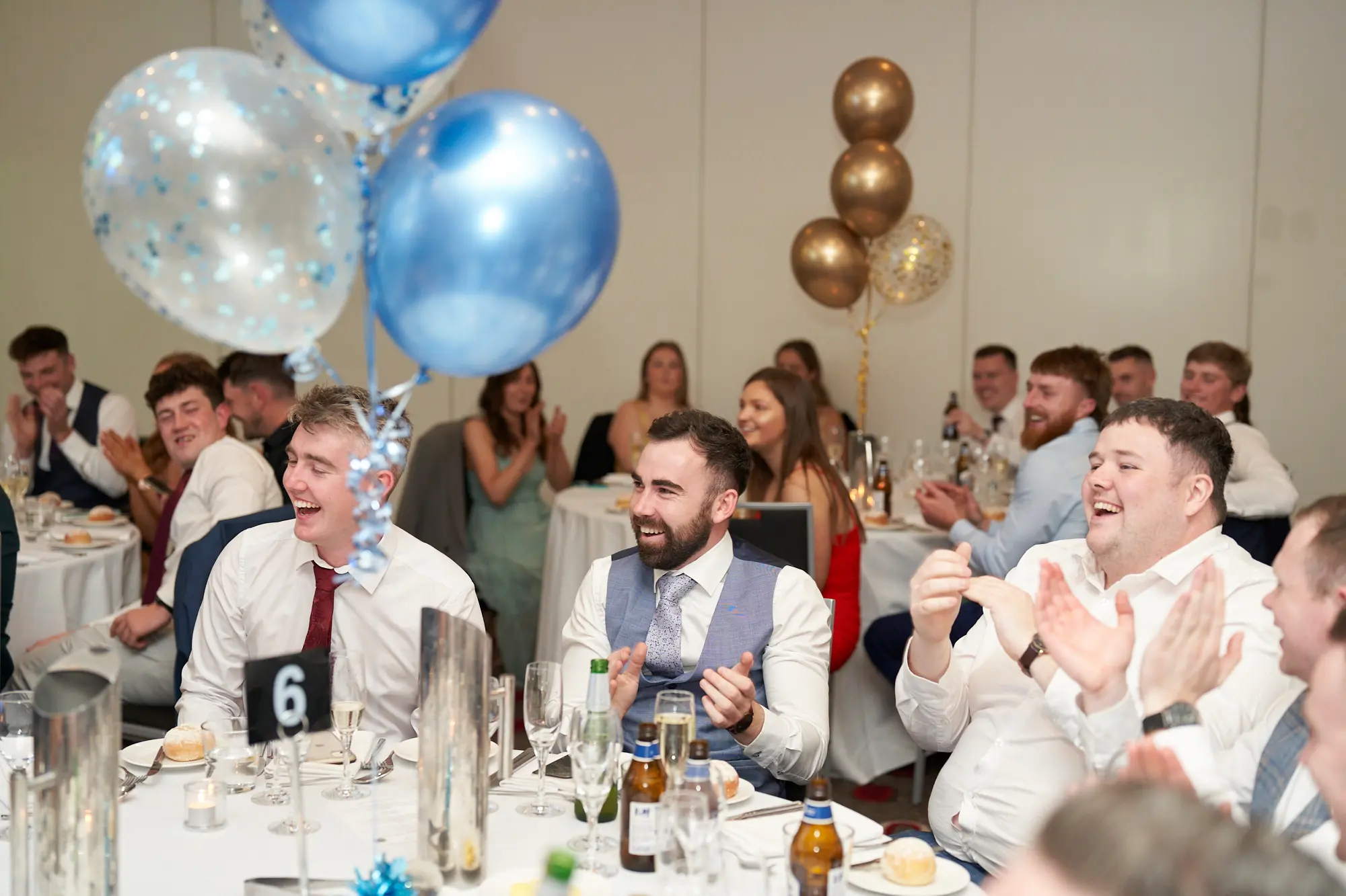 st kevins awards night pullman melbourne albert park candid lively clapping A candid moment captures three men at a dinner table reacting with laughter and applause during St. Kevin's Awards Night. The man on the right claps and laughs heartily, the center man in a waistcoat smiles broadly, and the man on the left in a white shirt and red tie looks toward the room with amusement. Guests at surrounding tables join in the celebration. The room features white tablecloths, formal place settings, and festive balloon displays in blue, gold, and confetti, conveying a lively, joyous, and celebratory awards night atmosphere.