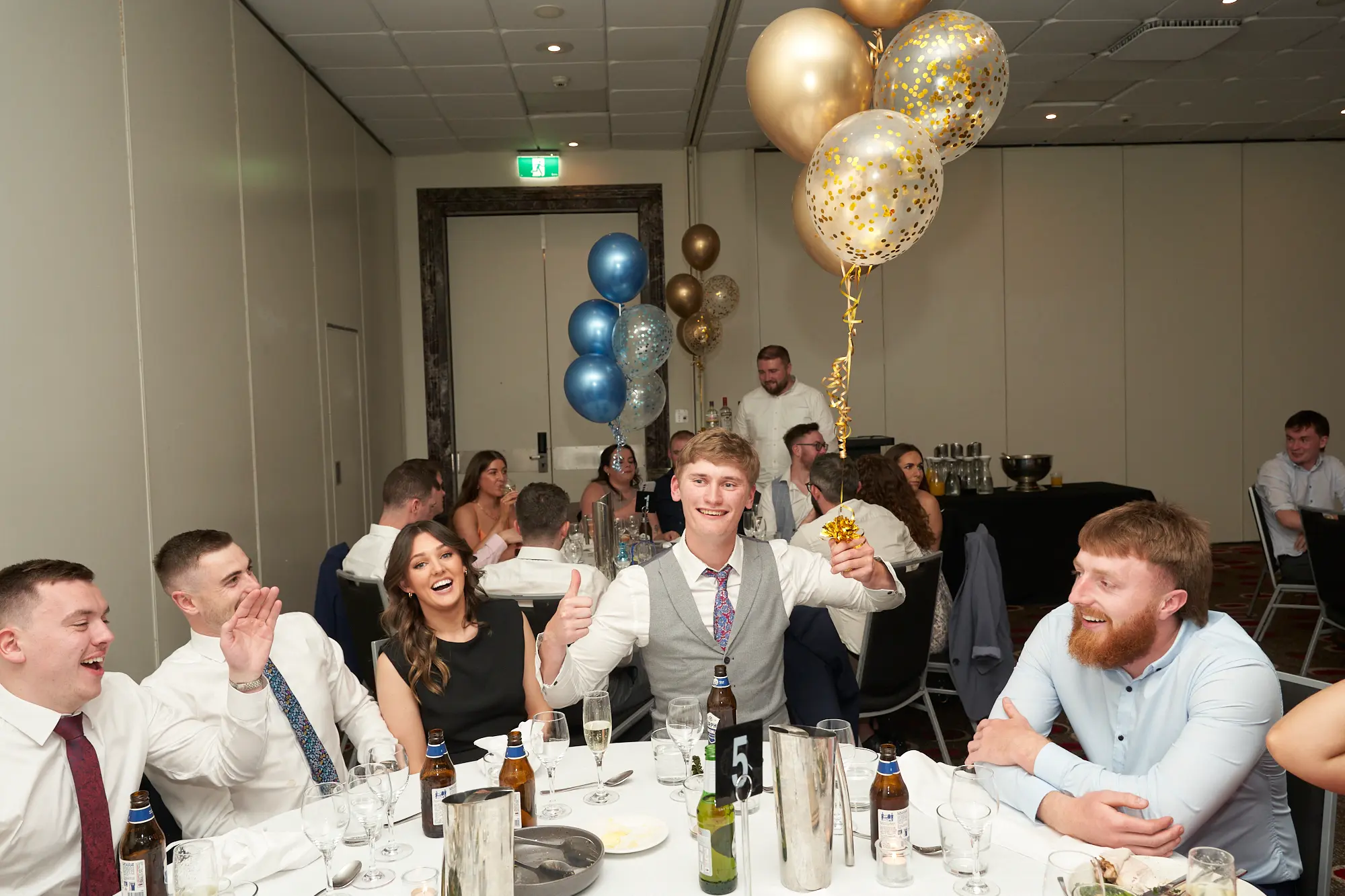 st kevins awards night pullman melbourne albert park candid thumbs up celebration A candid photo captures a lively group of young adults at a round dinner table during St. Kevin's Awards Night. A blonde man in a waistcoat smiles broadly at the camera, giving two thumbs up, while a woman in black laughs heartily beside him. Other guests at the table smile and chat, enjoying the evening. The table is formally set with plates, glassware, cutlery, and beer bottles, with gold and confetti balloons adding festive flair. The candid expressions, playful gestures, and elegant dinner setting convey the joy and celebratory energy of the awards night.