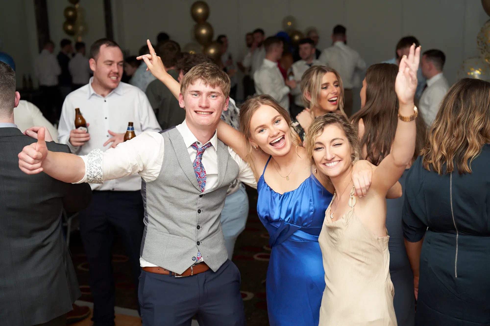 st kevins awards night pullman melbourne albert park celebration cheers This wide shot captures a lively group of friends on the St. Kevin's Awards Night dance floor. A man in a grey waistcoat smiles broadly with arms around two women—one in royal blue laughing exuberantly, the other in beige raising her hands in celebration. Nearby, a man in a white shirt gives a thumbs-up while holding a beer, and another woman laughs heartily. Surrounded by guests mingling and festive gold and blue balloons overhead, the scene radiates joyful energy, camaraderie, and the vibrant atmosphere of a post-awards party filled with laughter, cheers, and lively celebration.