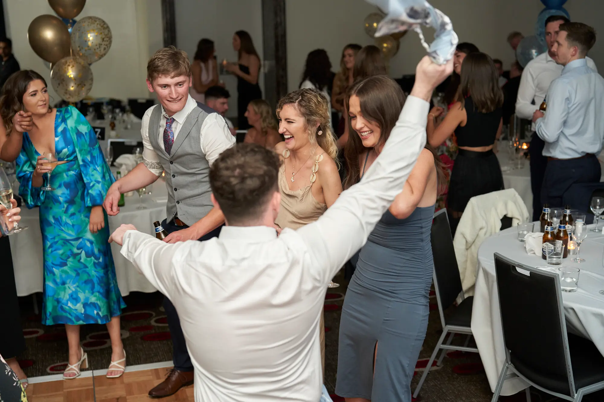 st kevins awards night pullman melbourne albert park celebration energy This wide shot captures a dynamic moment on the dance floor at the St. Kevin's Awards Night. A man in a white shirt raises his arms enthusiastically, holding a white handkerchief, while guests around him laugh, smile, and interact. Women in vibrant floral and gold satin dresses, and a man in a grey waistcoat, are captured mid-celebration. Festive gold, blue, and confetti balloons hover above, and round dinner tables with drinks and glassware fill the background. The image conveys lively engagement, spontaneous joy, and the energetic, social atmosphere of a post-awards party.