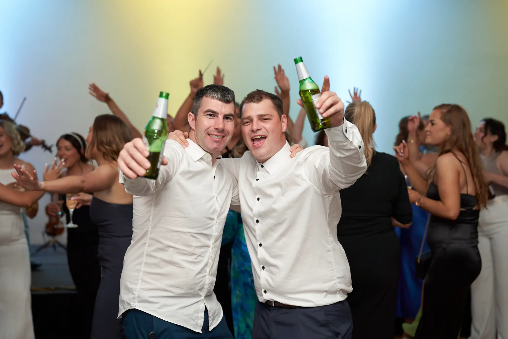 st kevins awards night pullman melbourne albert park cheerful toast dance floor This wide shot captures a lively moment on the dance floor at the St. Kevin's Awards Night. Two young men in white shirts and dark trousers pose enthusiastically, arms around each other, holding green beer bottles in a celebratory toast. Behind them, guests dance with hands in the air, laugh, and mingle, while a live band with a violinist performs on stage. The wooden dance floor, warm ambient and blue stage lighting, and vibrant energy highlight the joyous, social, and festive atmosphere of the post-awards celebration.