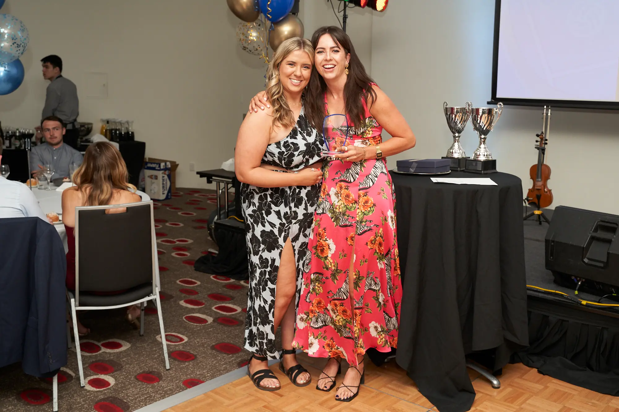 st kevins awards night pullman melbourne albert park coral pink award stage Two women pose on stage at St. Kevin's GAC 2023 Awards Night at Pullman Melbourne Albert Park. The woman on the left wears a black-and-white floral one-shoulder dress, smiling with her arm around the woman on the right, who wears a vibrant coral-pink dress with a zebra pattern and holds a clear glass award plaque. The stage features balloons, silver trophies, a violin, projection screen, and lighting, creating a celebratory and formal atmosphere.