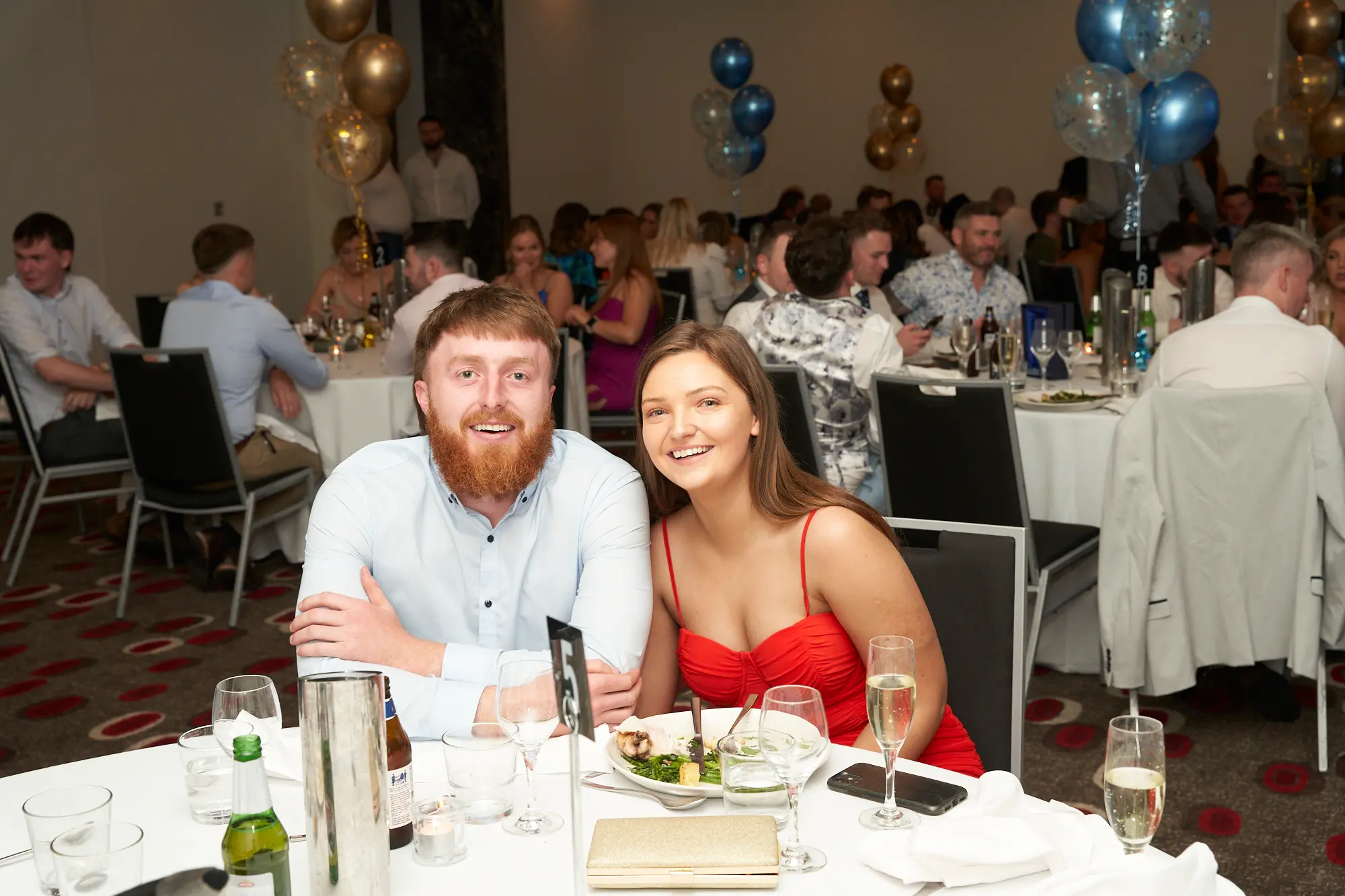 st kevins awards night pullman melbourne albert park couple smiling table number 5 A man in a light blue shirt and a woman in a red spaghetti-strap dress smile brightly for the camera while seated together at a dinner table during St. Kevin’s Awards Night at Pullman Melbourne Albert Park. Plates of food, glasses, and a table number “5” are visible in front of them. In the background, guests dressed in formal attire are seated and mingling at round tables, surrounded by festive blue and gold balloons. The cheerful expressions and elegant setting capture the warm and joyful spirit of the evening celebration.