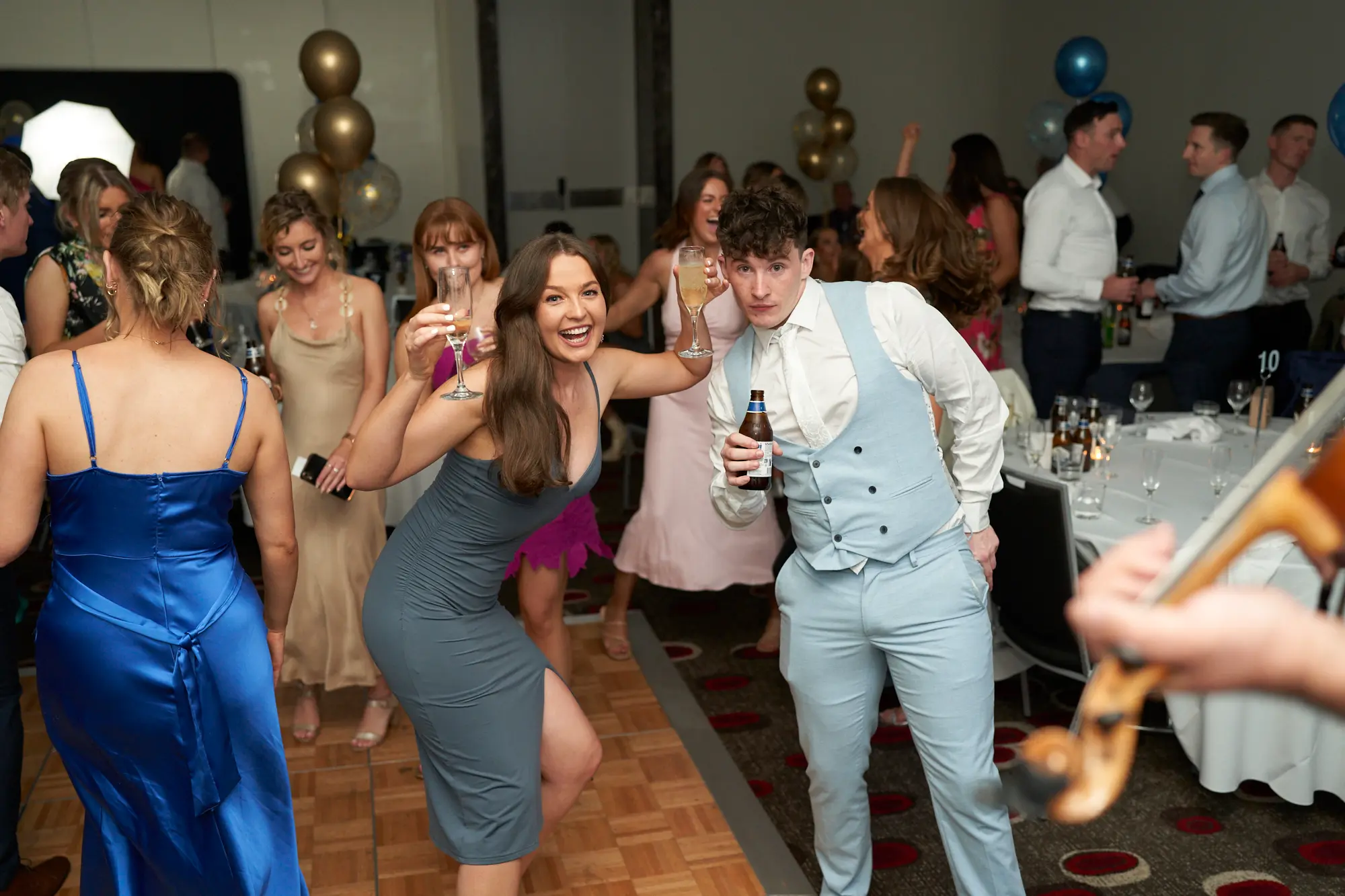 st kevins awards night pullman melbourne albert park dance floor celebration candid moments A joyful, high-energy scene unfolds on the dance floor during St. Kevin’s Awards Night at Pullman Melbourne Albert Park. A woman in a grey bodycon dress with a side slit crouches playfully, smiling at the camera while holding two champagne flutes. Beside her, a man in a light blue three-piece suit holds a beer bottle, beaming proudly. Around them, guests in elegant evening wear dance, laugh, and mingle under soft, warm lighting. Gold and blue balloons and a glimpse of a live band’s violin add to the festive, vibrant atmosphere of the celebration.