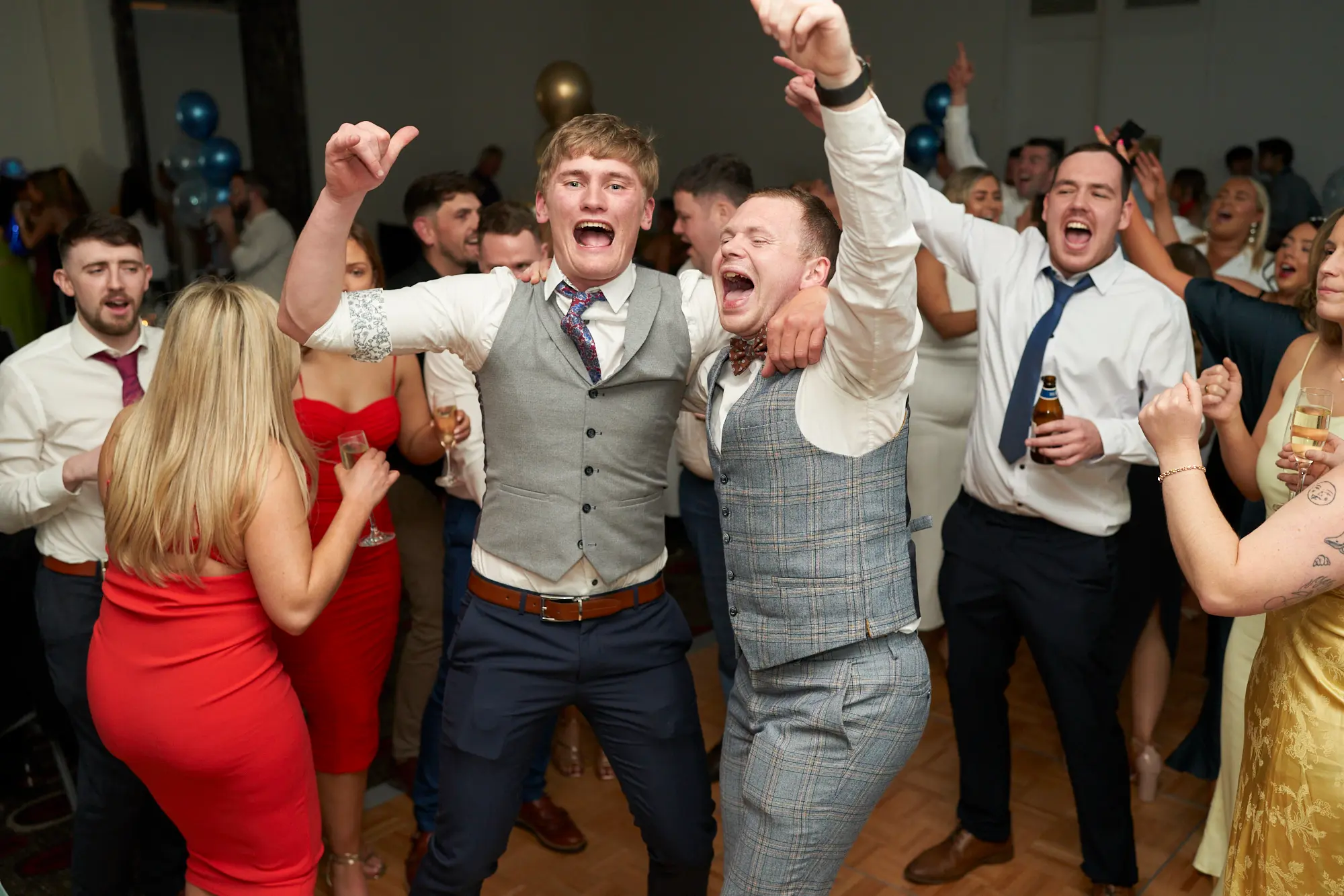 st kevins awards night pullman melbourne albert park dance floor celebration energy On the wooden parquet dance floor of St. Kevin's Awards Night, two young men are caught in a moment of exuberant celebration. The man in a grey waistcoat and dark trousers raises both hands, yelling or singing with joy, while his friend in a grey checkered three-piece suit wraps an arm around him, equally thrilled. Around them, formally dressed guests laugh, dance, and raise drinks. Warm ambient lighting and clusters of gold and blue balloons create a festive, energetic atmosphere, capturing the spirited camaraderie and high energy of a lively post-awards night party.