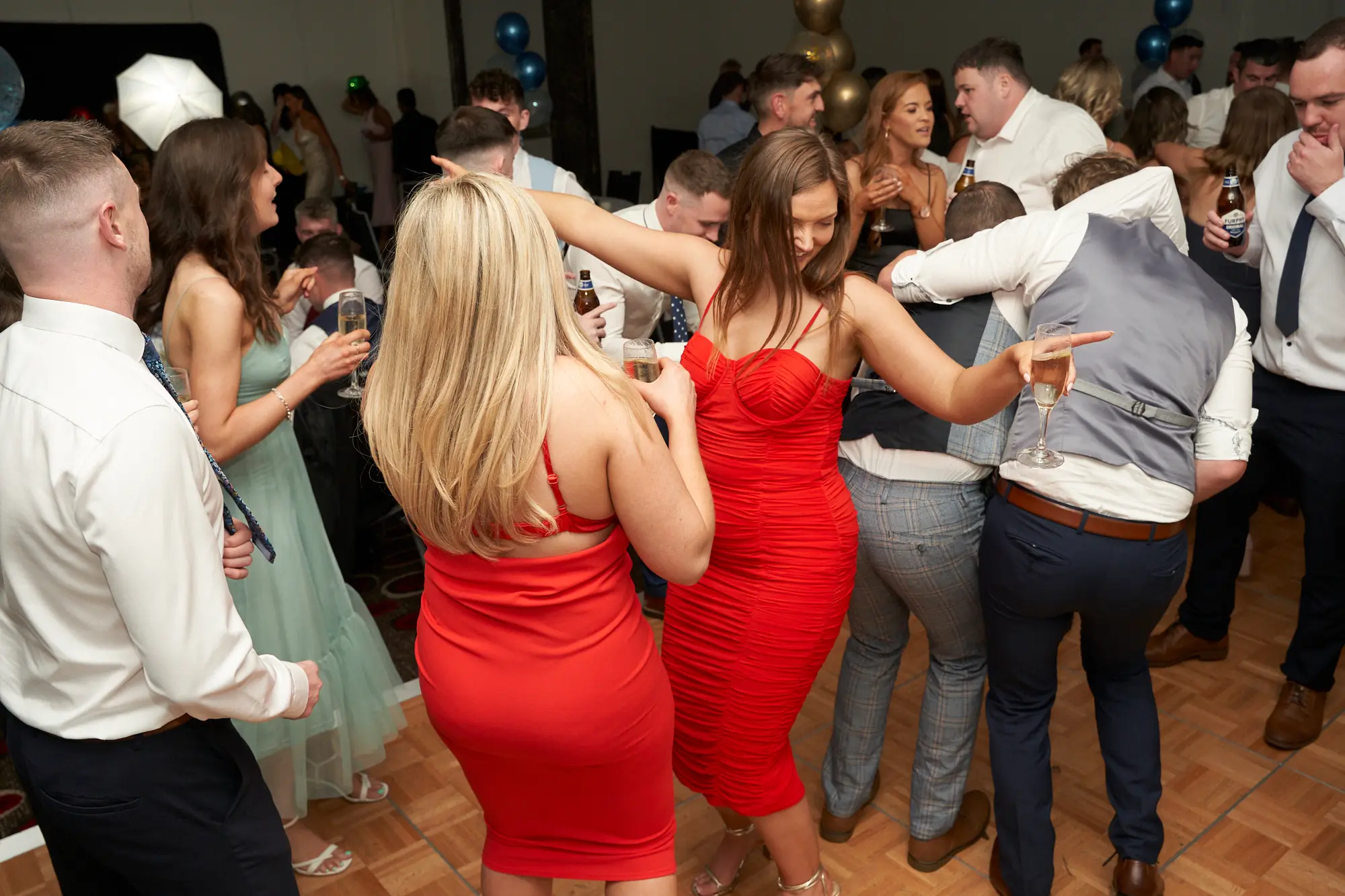 st kevins awards night pullman melbourne albert park dance floor celebration Guests celebrate on the dance floor during St. Kevin's Awards Night, with two women in vibrant red bodycon dresses dancing at the center, surrounded by friends smiling, chatting, and holding drinks. The wooden parquet floor and festive gold and blue balloons create a lively backdrop, while formally dressed attendees mingle, dance, and enjoy the energetic, joyful, and celebratory atmosphere of the post-awards party.