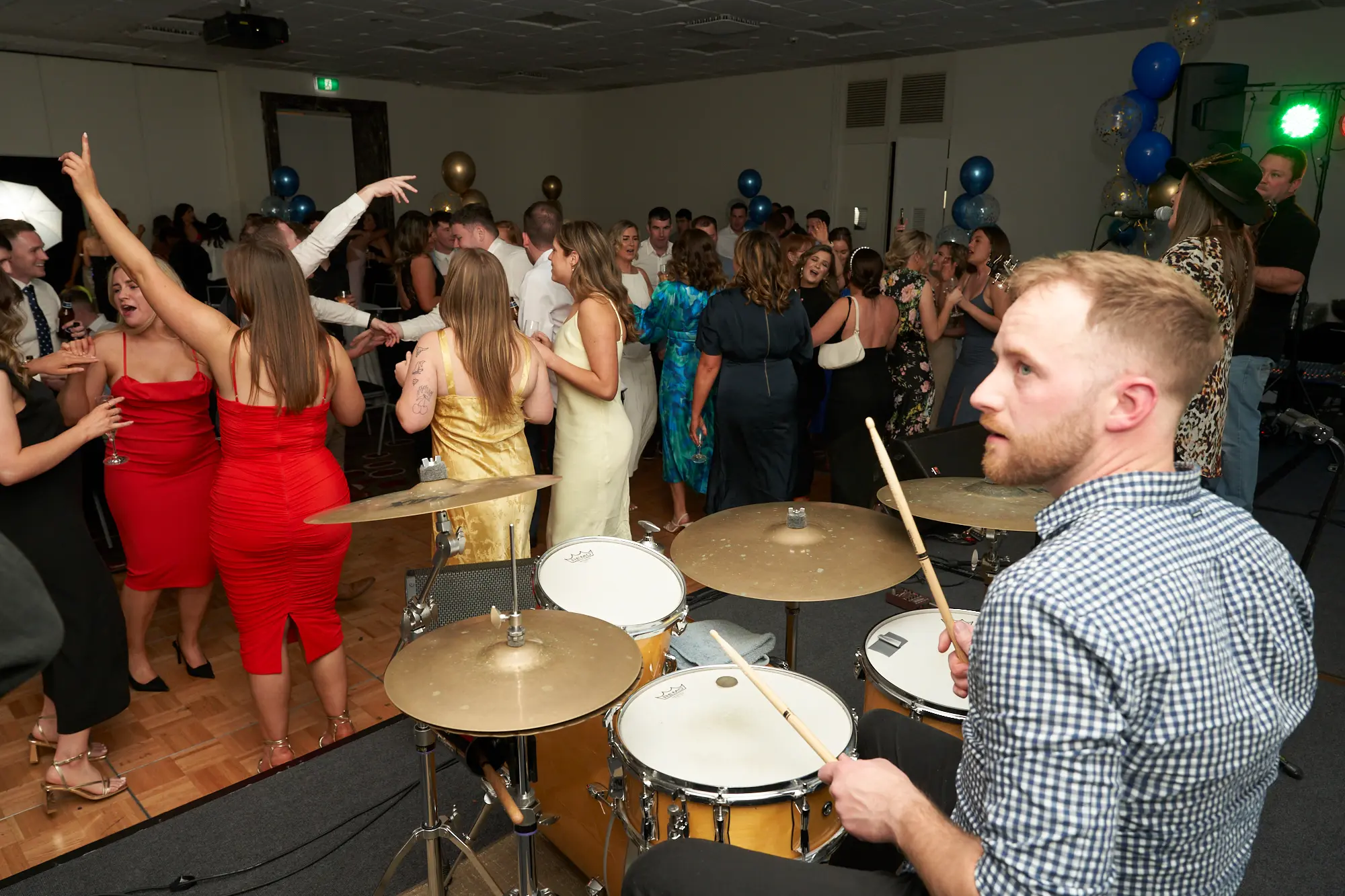 st kevins awards night pullman melbourne albert park dance floor drummer guest celebration A lively scene on the dance floor at St. Kevin's Awards Night captures the energy of the celebration. In the foreground, a man in a blue and white checkered shirt sits behind a natural wood-finish drum set, holding drumsticks and focused on playing. Behind him, formally dressed guests mingle, dance, and socialize. A woman in a vibrant red dress raises her arm in joy, smiling broadly. The room is decorated with clusters of gold and blue balloons, and a warm, festive atmosphere fills the space, reflecting the excitement and camaraderie of the post-awards party.