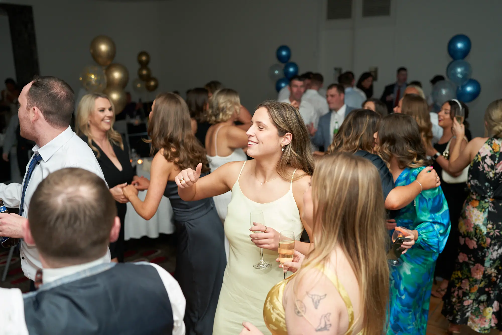 st kevins awards night pullman melbourne albert park dance floor energy Guests celebrate on the dance floor during St. Kevin's Awards Night, with a woman in a light yellow slip dress laughing and holding a champagne flute, surrounded by friends smiling and engaging. Festive gold, blue, and confetti balloons decorate the room, while formally dressed attendees mingle, dance, and socialize. The dark patterned carpet and neutral walls frame the lively, energetic, and joyful atmosphere, capturing the spirited post-awards party as guests enjoy music, conversation, and movement across the floor.