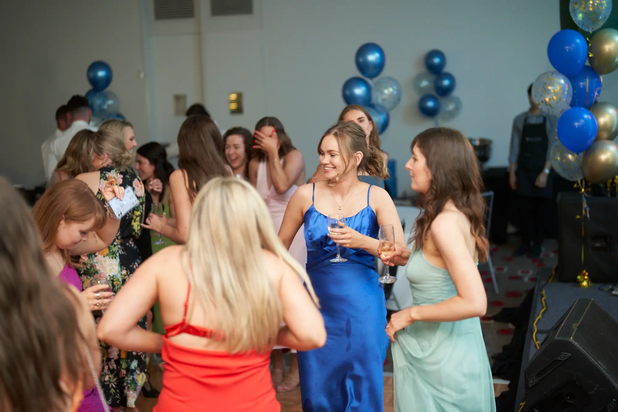 st kevins awards night pullman melbourne albert park dance floor laughter This wide shot captures a lively dance floor moment at the St. Kevin's Awards Night. In the center, a woman in a royal blue satin slip dress and a woman in a mint-green slip dress smile and toast with champagne flutes. Around them, other women dance, laugh, and interact enthusiastically. Festive gold, blue, and confetti balloons decorate the room, while a large black speaker and other event equipment are visible in the background. The image conveys vibrant energy, camaraderie, and joy, highlighting the celebratory and dynamic atmosphere of the post-awards party.