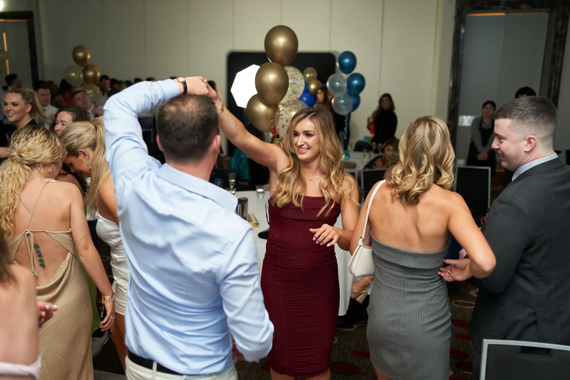 st kevins awards night pullman melbourne albert park dance floor maroon smiles This wide shot captures a lively, energetic moment on the dance floor at the St. Kevin's Awards Night. A woman in a vibrant maroon slip dress smiles broadly at the camera while holding a champagne flute. To her left, a man in a light blue shirt leads a partner in a dance move, while a woman in a strapless grey dress holds a white handbag. Guests in formal attire mingle, laugh, and socialize around decorated tables with festive balloons. The dark patterned carpet and celebratory atmosphere reflect the joyful energy of a post-awards party.