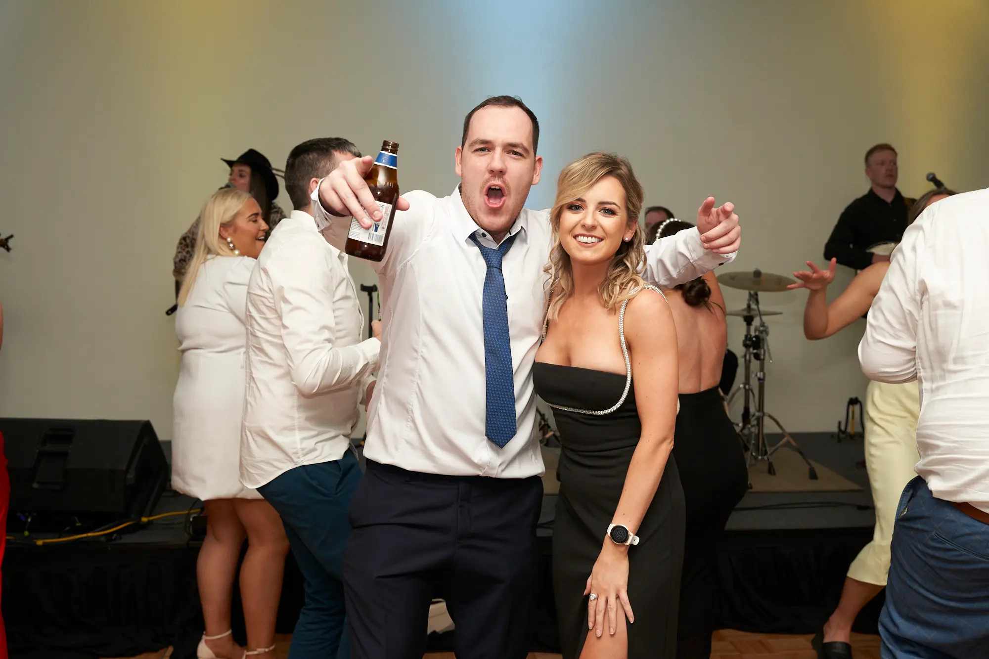 st kevins awards night pullman melbourne albert park dance floor spotlight This wide shot captures a high-energy moment on the dance floor during the St. Kevin's Awards Night. A man in a white shirt and blue patterned tie points enthusiastically at the camera, holding a beer, while a blonde woman in a black strapless dress mirrors his gesture with a bright smile. Guests around them laugh, dance, and socialize, including a woman in a white dress laughing heartily. A live band with a violinist and drummer performs in the background, with stage lights and speakers adding to the festive, celebratory atmosphere of this post-awards party.