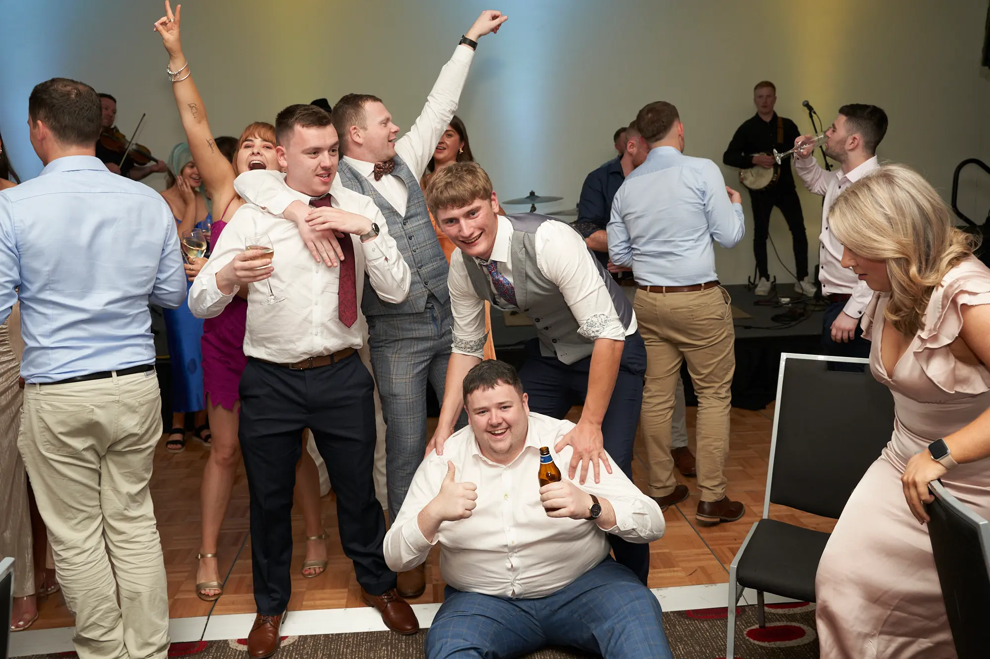 st kevins awards night pullman melbourne albert park dance floor thumbs up This wide shot captures a lively moment on the dance floor at the St. Kevin's Awards Night. A bearded man in a white shirt sits in the foreground, smiling widely and giving a double thumbs-up while holding a beer bottle. Around him, guests pose enthusiastically, including a man in a grey checkered waistcoat, a man in a white shirt with a tie, and a woman in a red dress flashing a peace sign. Live musicians perform in the background, with violin, acoustic guitar, banjo, and drums, while festive balloons and stage lighting enhance the celebratory atmosphere.