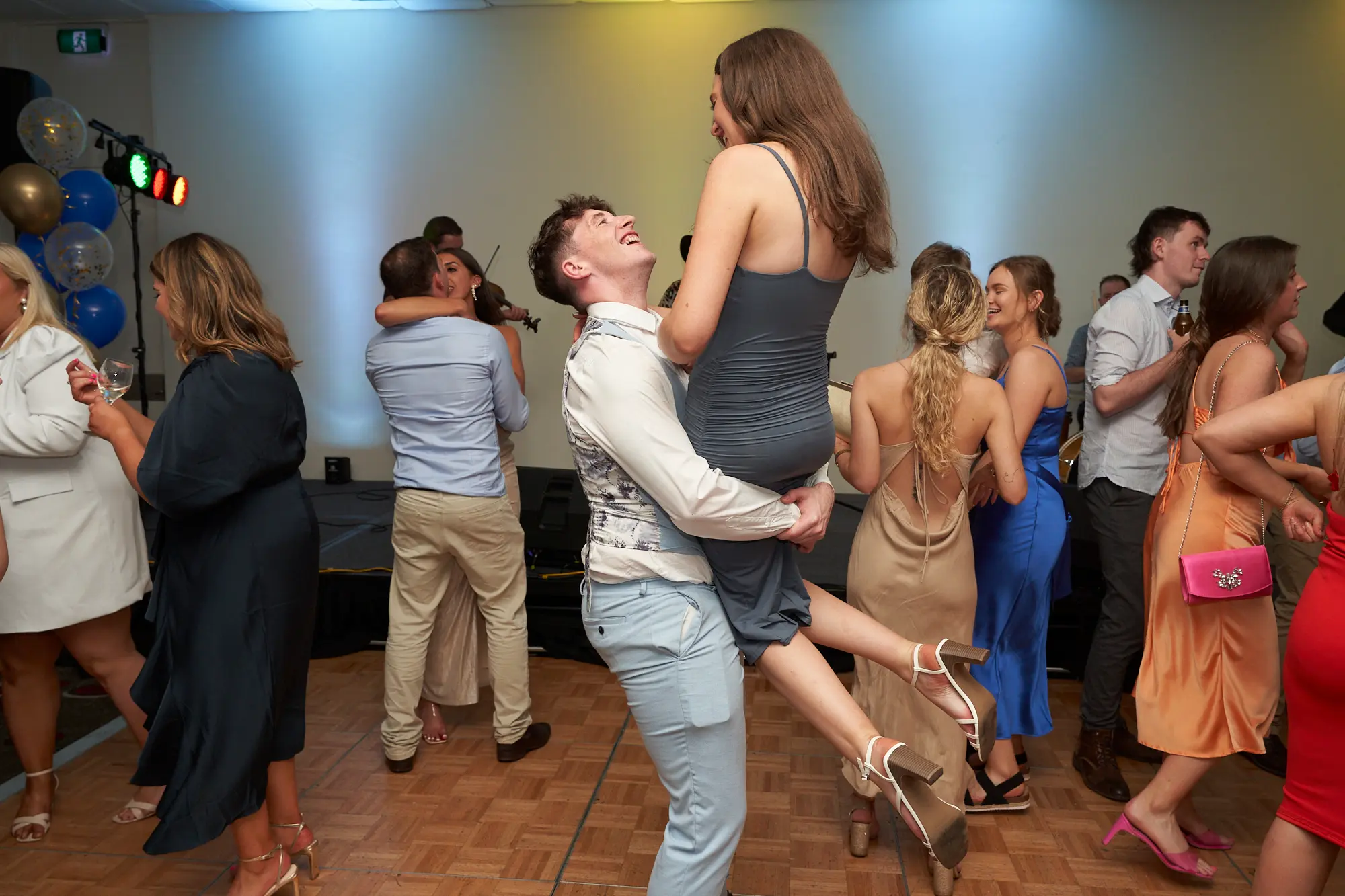 st kevins awards night pullman melbourne albert park dance lift This wide shot captures a joyful couple on the dance floor at the St. Kevin's Awards Night. The man, in a white shirt and light blue trousers, dips and lifts a woman in a dark grey slip dress and white heels, both laughing and locked in the moment. Around them, guests in formal attire dance, mingle, and hold drinks, with gold and blue balloons adding a festive touch. Stage lighting washes the room in warm yellow and cool blue, highlighting the lively energy, laughter, and celebratory atmosphere of the post-awards party.