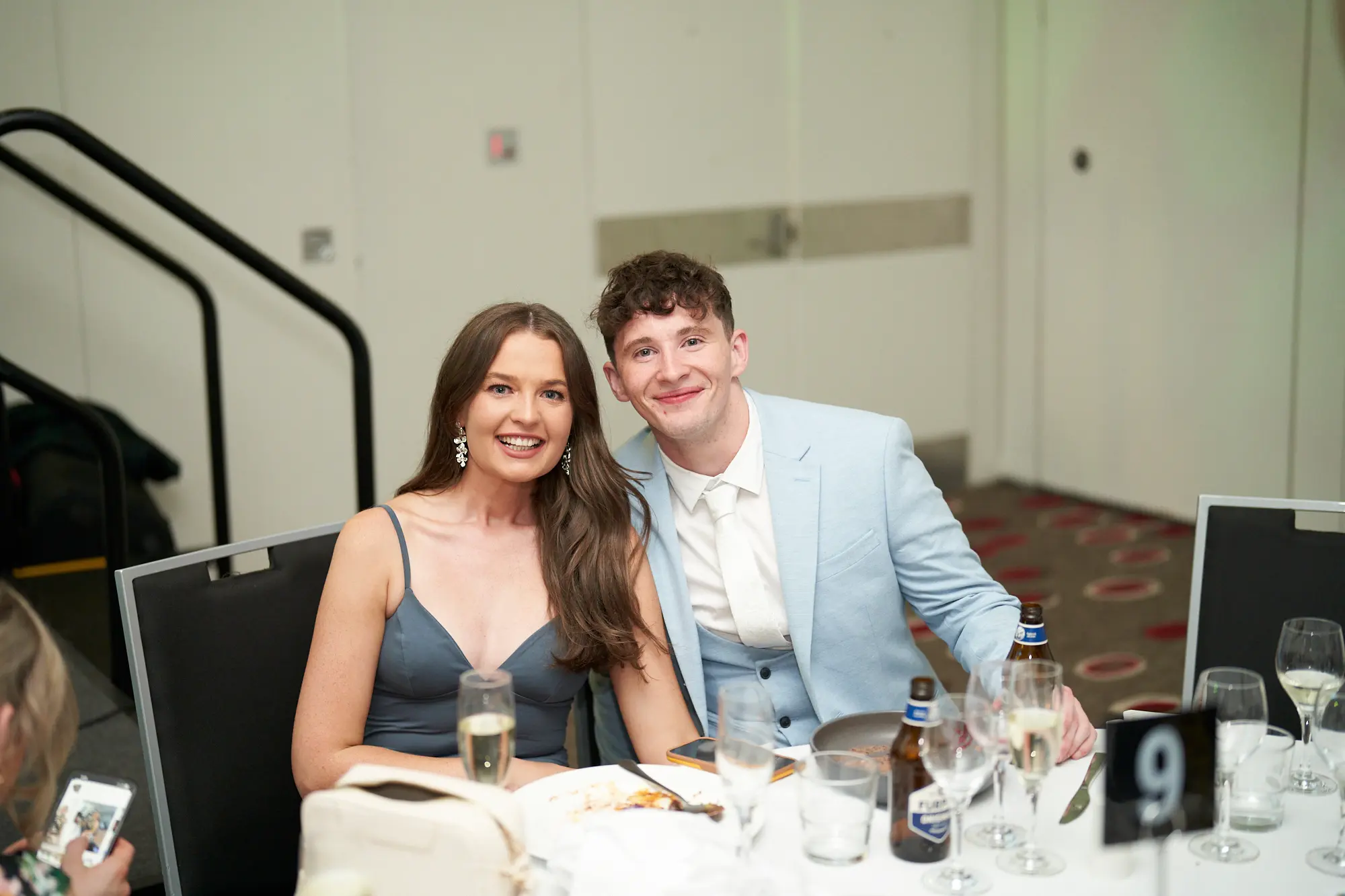 st kevins awards night pullman melbourne albert park dinner pose man woman A man and a woman pose together at a dinner table during the St. Kevin's Awards Night. The woman, wearing a dark blue-grey v-neck slip dress with long drop earrings, holds a champagne flute and smiles warmly. The man, in a light blue three-piece suit with a white tie, smiles broadly beside her. The table is set with glassware, cutlery, plates, and two beer bottles, with a table number "9" card visible. The indoor function room features light beige walls, a dark patterned carpet, and a social, celebratory atmosphere as guests enjoy the evening.