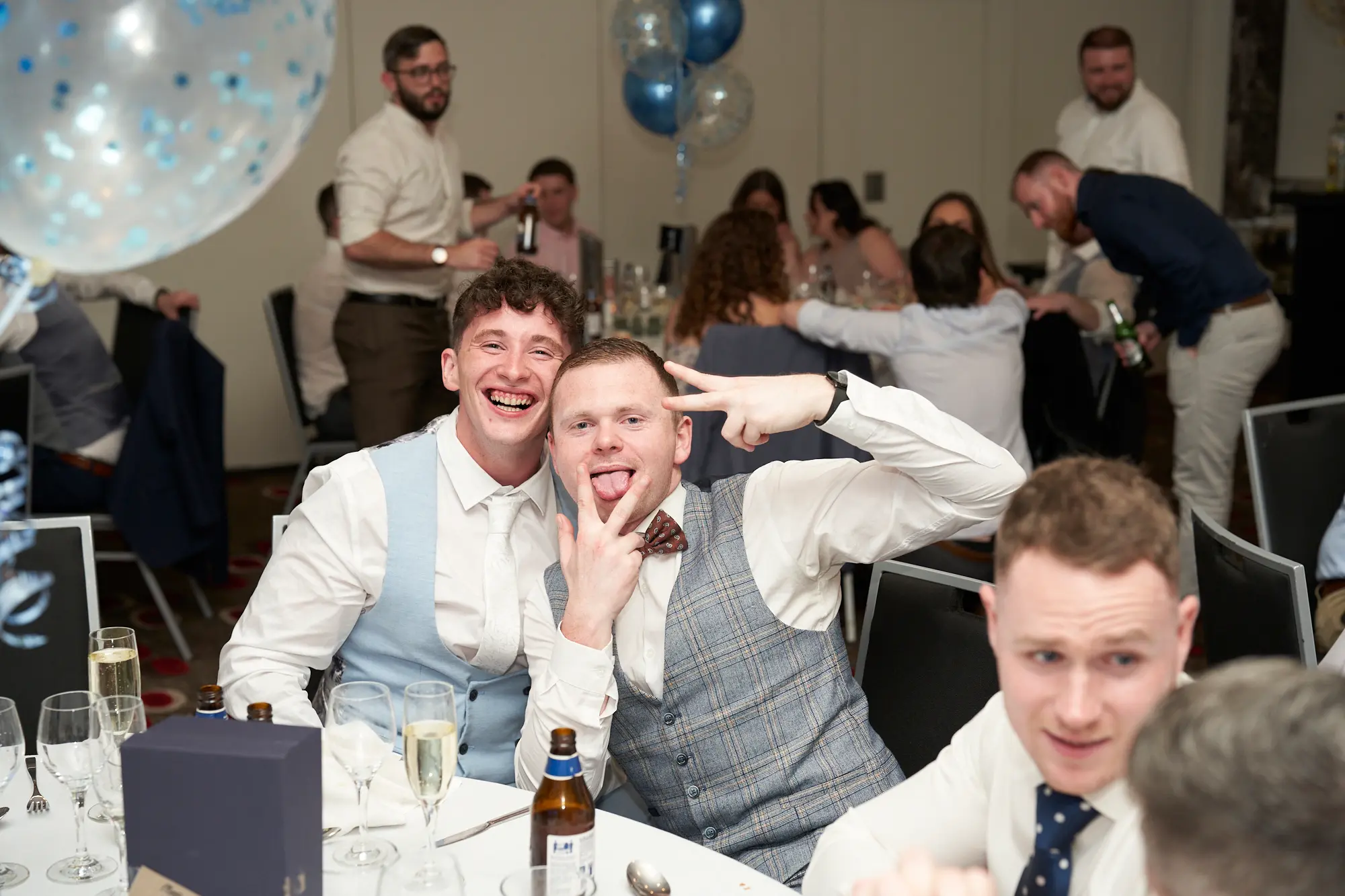 st kevins awards night pullman melbourne albert park dinner table candid laughter Two young men share a playful moment during the St. Kevin's Awards Night dinner, seated at a white-clothed table. The man on the left wears a light blue waistcoat and white shirt, laughing with his mouth open wide, while the man on the right wears a grey checkered waistcoat and patterned bowtie, sticking his tongue out and flashing a double peace sign. Behind them, other guests in formal attire mingle and hold drinks, with large clusters of gold, blue, and clear confetti balloons adding to the festive indoor setting. The scene captures joyful, candid celebration among attendees.