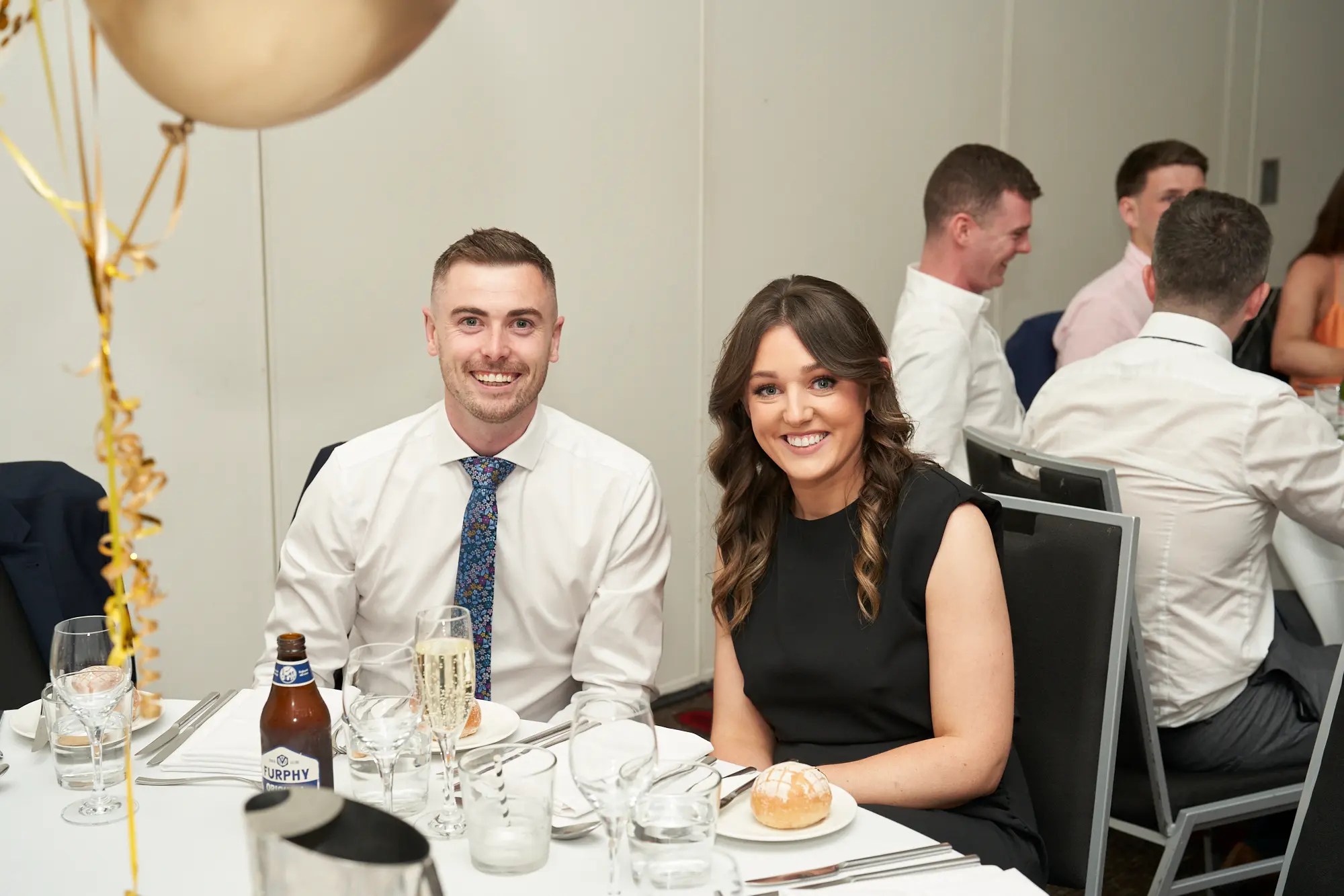 st kevins awards night pullman melbourne albert park dinner table guest portrait Two guests are seated together at a formally set dinner table during the annual St. Kevin's Awards Night, smiling warmly at the camera. The man on the left wears a white button-up shirt and blue patterned tie, while the woman on the right wears a black sleeveless dress. A bread roll and a beer bottle labeled “FURPHY” are on the table. Festive gold and clear confetti balloons decorate the room, and other formally dressed guests are visible at nearby tables. The candid, joyful expressions and elegant setting convey a relaxed, celebratory awards night atmosphere.