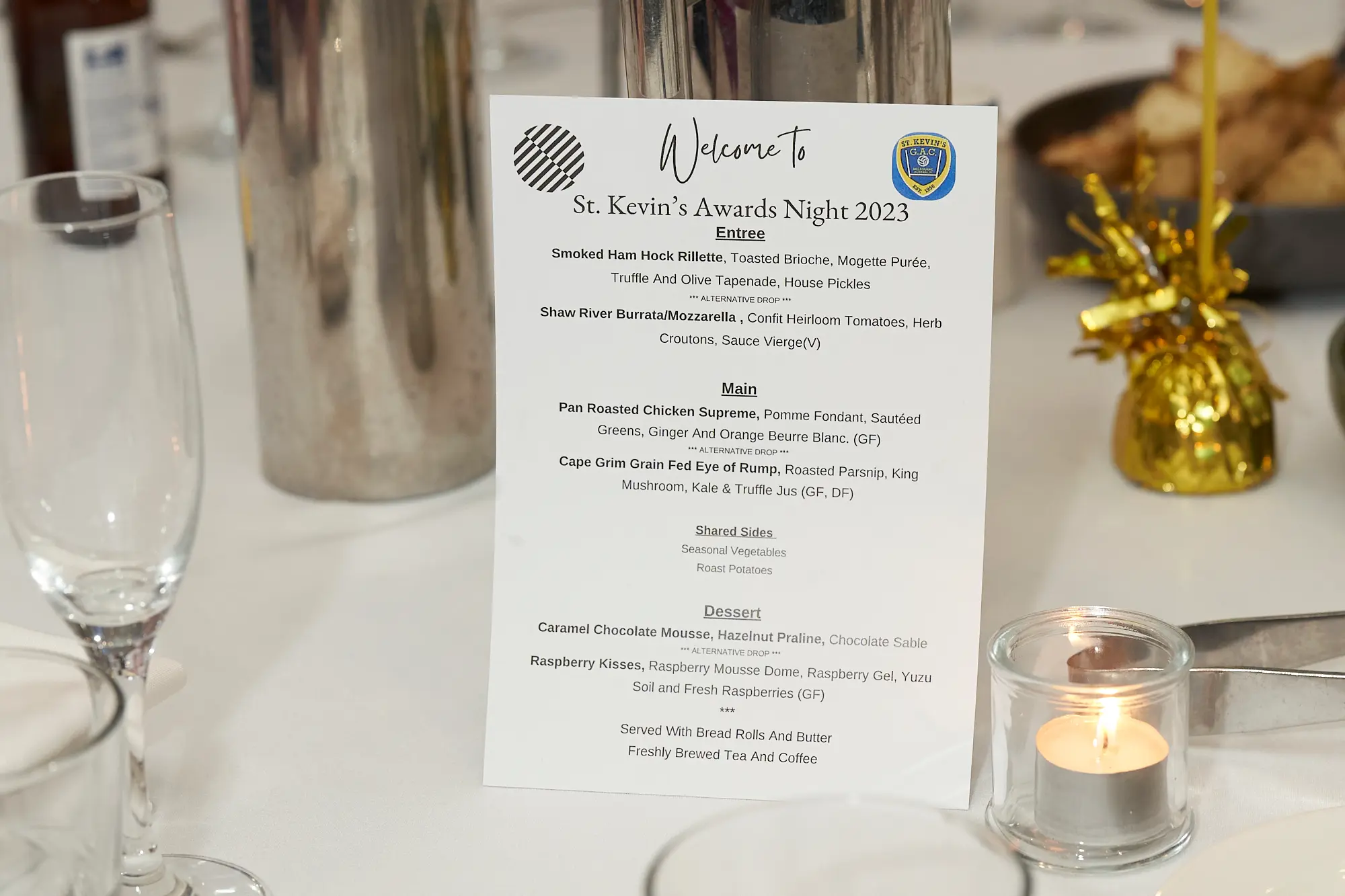 st kevins awards night pullman melbourne albert park dinner table menu setting A beautifully arranged dinner table at St. Kevin's Awards Night at Pullman Melbourne Albert Park showcases a printed menu card for the 2023 event. The menu lists gourmet selections, including smoked ham hock rillette, pan roasted chicken supreme, and caramel chocolate mousse. Wine glasses, a glowing candle in a glass holder, and gold-colored table decorations create an elegant dining atmosphere. The menu card features two logos — a stylized wavy circle and the St. Kevin’s GAC emblem — reflecting a refined and festive setup for this special awards night celebration.