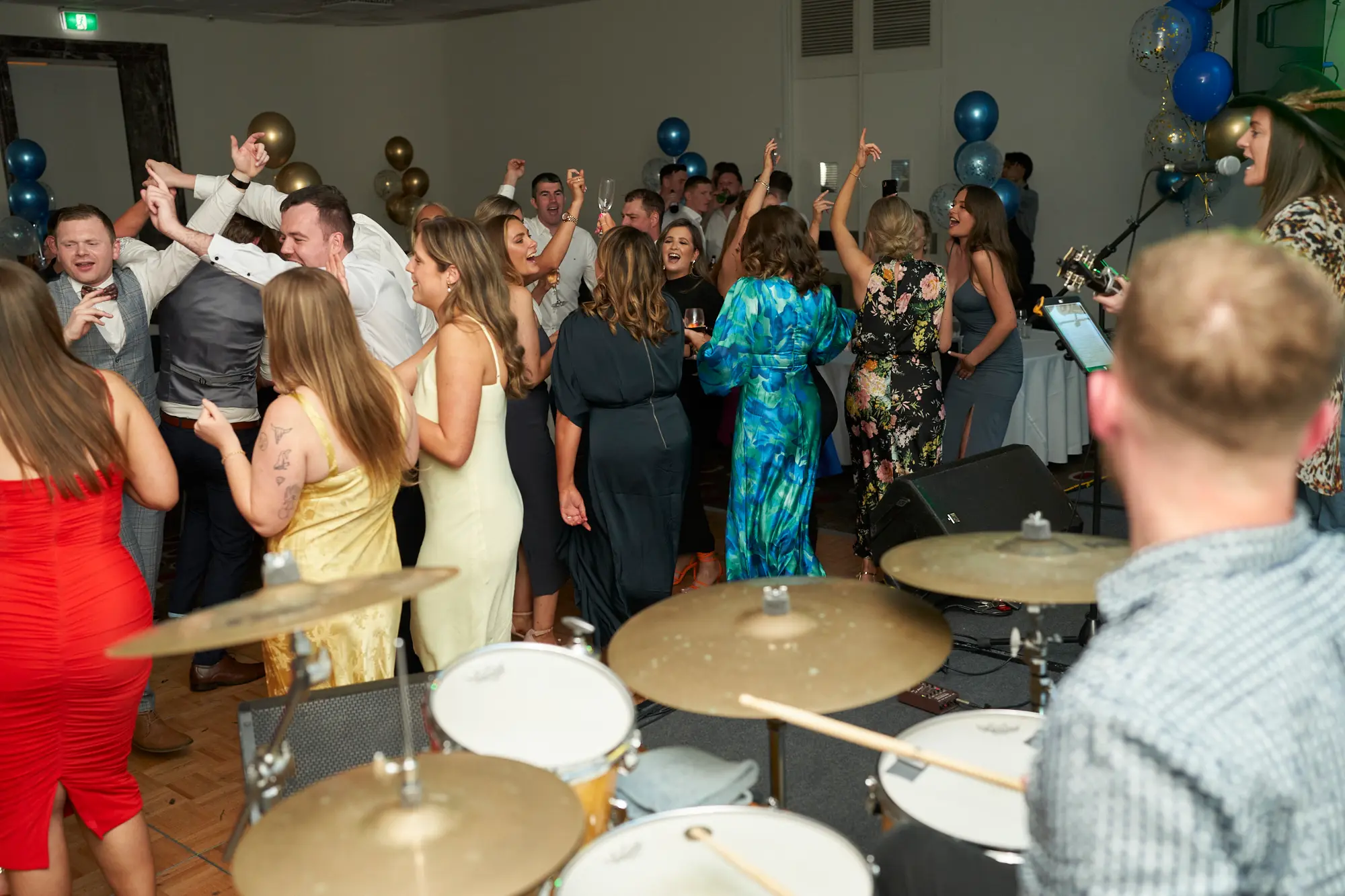 st kevins awards night pullman melbourne albert park drummer perspective energy This wide shot captures a high-energy moment on the dance floor at the St. Kevin's Awards Night, viewed from just behind the drummer. Guests in formal evening wear are dancing, laughing, and holding drinks, with a man in a dark waistcoat raising both hands enthusiastically. A woman in a vibrant blue floral dress holds a glass nearby, while others mingle and celebrate. Gold, blue, and confetti balloons, live band instruments, and formal dinner tables set the festive scene. The image conveys spontaneous joy, dynamic movement, and the lively spirit of a post-awards party celebration.