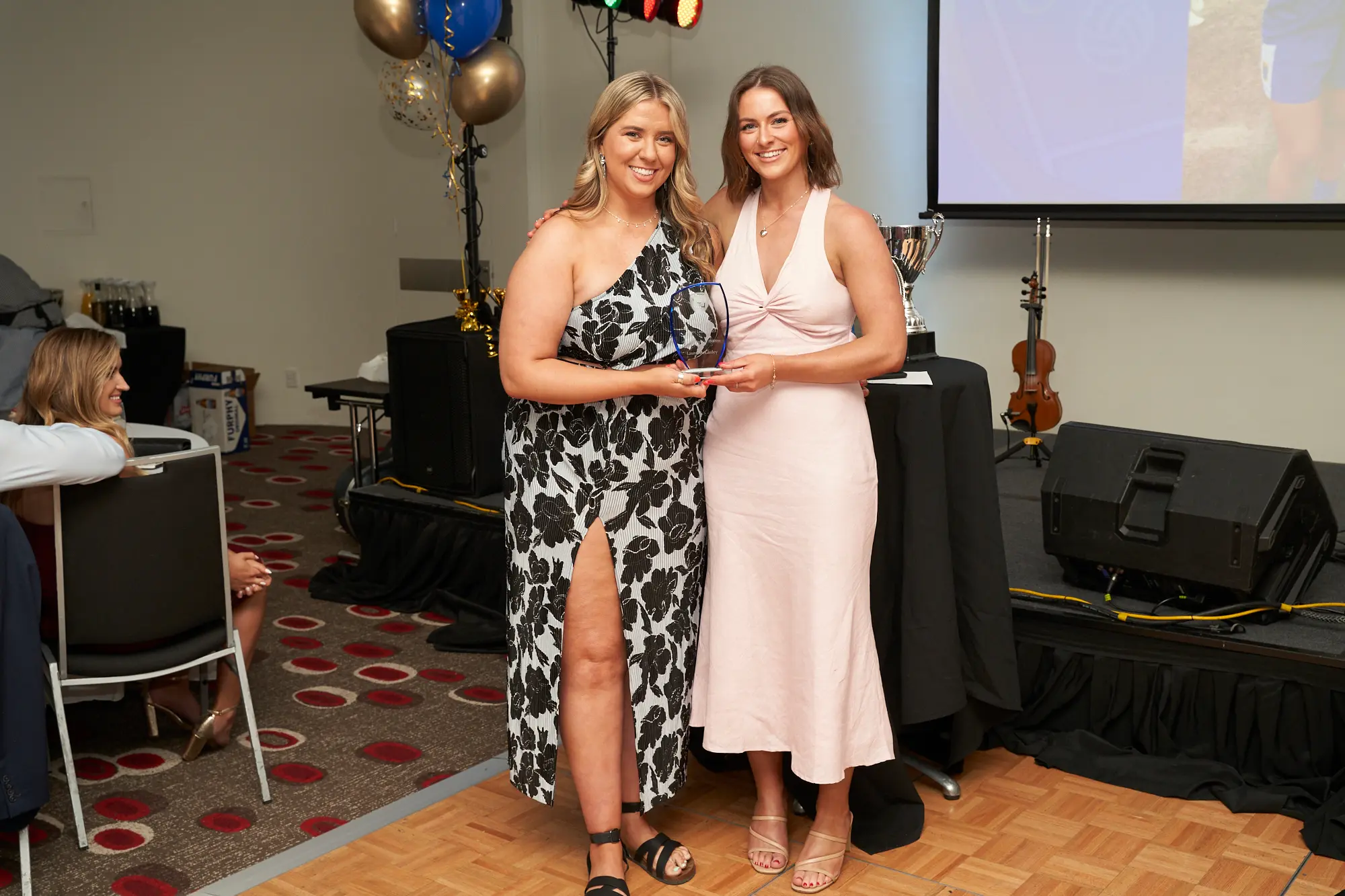 st kevins awards night pullman melbourne albert park floral award stage Two women pose on stage at St. Kevin's GAC 2023 Awards Night at Pullman Melbourne Albert Park. The woman on the left wears a black-and-white floral one-shoulder dress with a high slit, holding a clear glass award plaque and smiling. Beside her, a woman in a light pink sleeveless dress also smiles. The stage features balloons, silver trophies, a violin, and a partially visible projection screen, creating a celebratory and formal atmosphere.