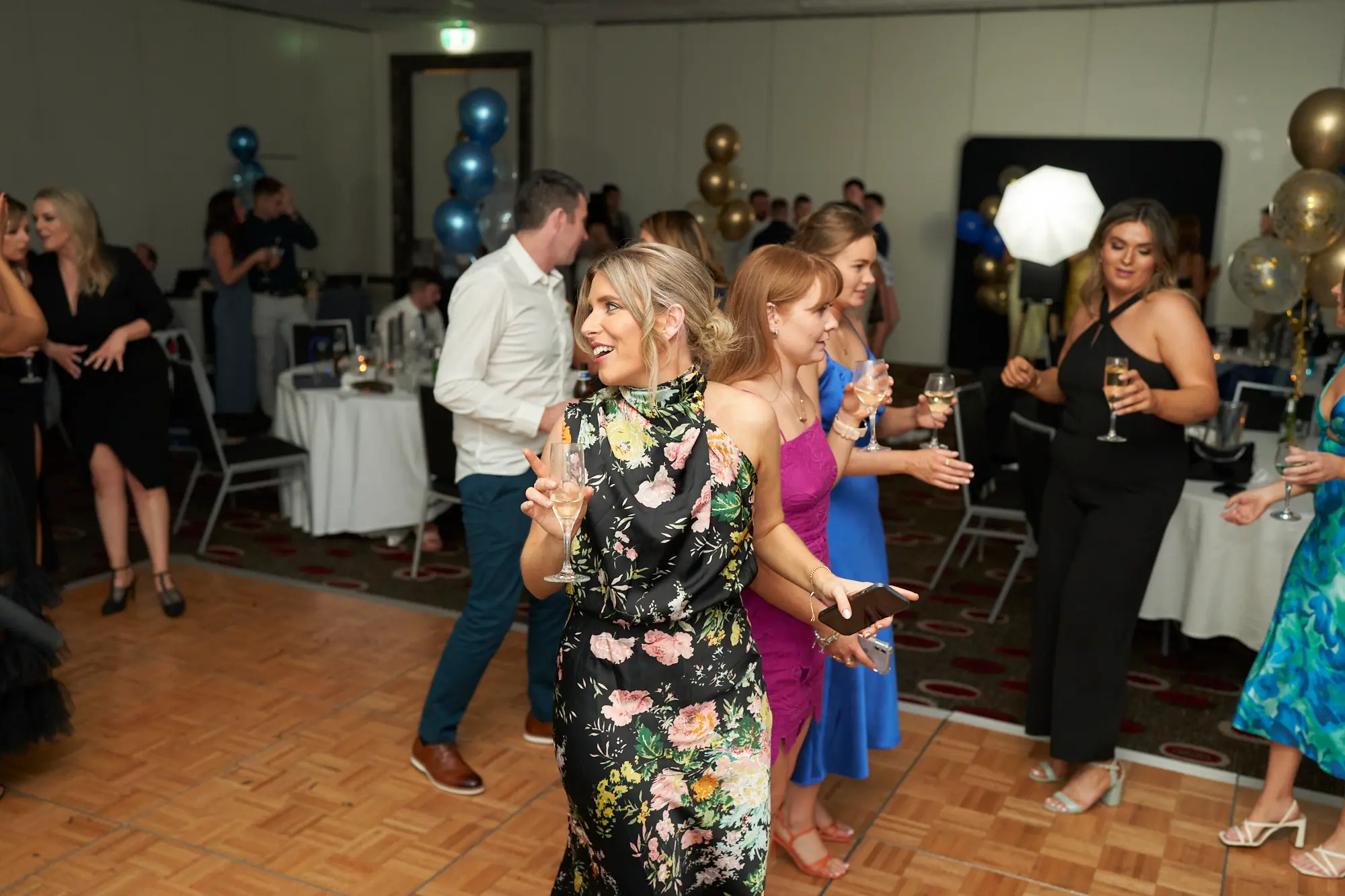 st kevins awards night pullman melbourne albert park floral laughter This wide shot captures a joyful, energetic moment on the dance floor at the St. Kevin's Awards Night. A blonde woman in a black floral dress laughs heartily, holding a wine glass, while friends in fuchsia, royal blue, and printed dresses join in the celebration with drinks in hand. Guests in the background mingle near dinner tables and a black stage backdrop featuring a glowing white sphere. Candid smiles, animated gestures, and festive gold and blue balloons above convey the lively, social, and celebratory atmosphere of a post-awards night party.