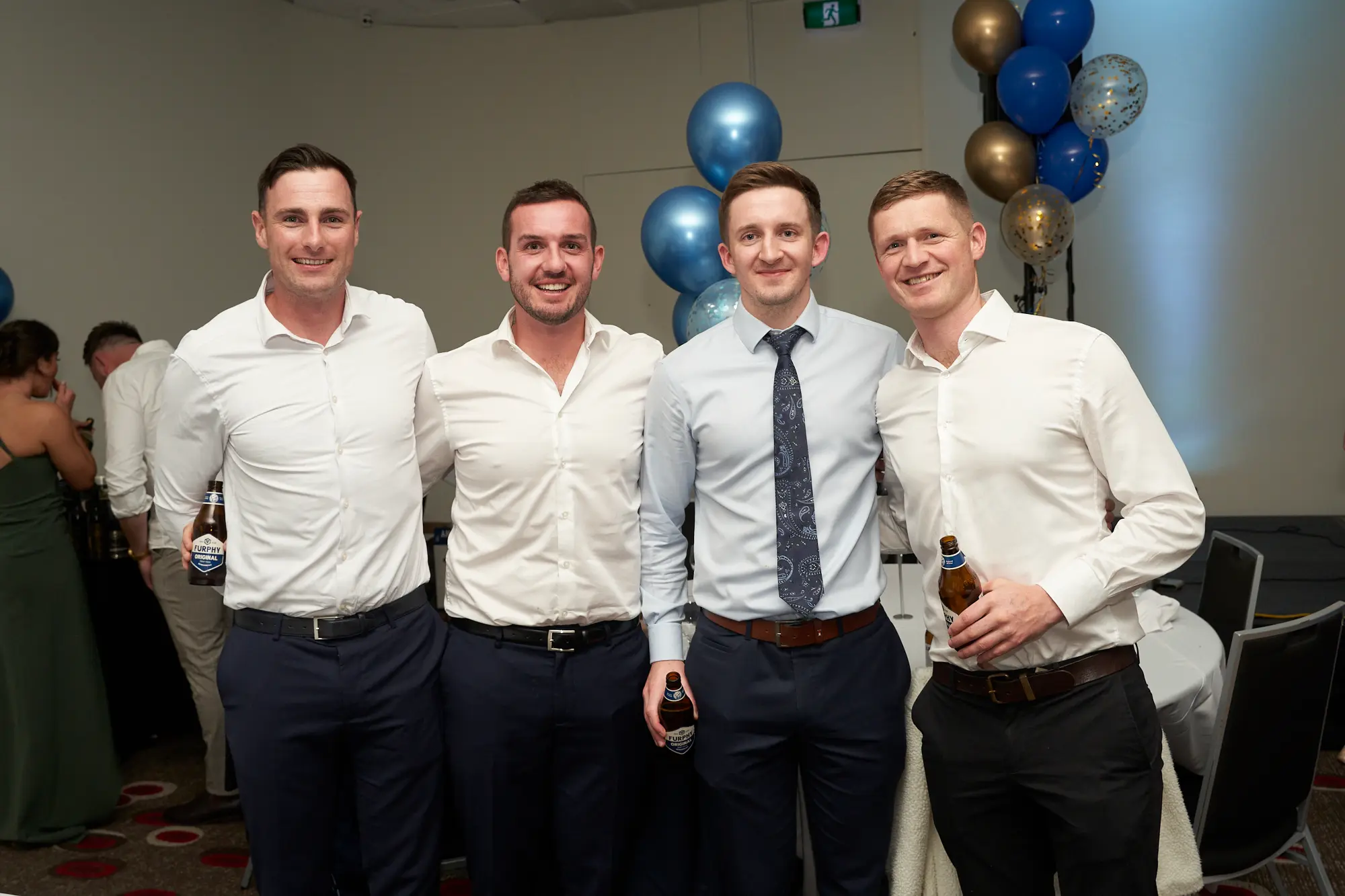st kevins awards night pullman melbourne albert park four men celebration Four men stand side-by-side at the St. Kevin's Awards Night, smiling warmly at the camera. They are dressed in white shirts and dark trousers or suits, three holding beer bottles, and one wearing a tie. Behind them, a festive display of blue and gold balloons adds color, while other attendees are visible near white-clothed tables. The indoor function room has plain beige walls and warm lighting. The men’s relaxed poses and cheerful expressions convey a social and celebratory atmosphere, capturing a joyful post-ceremony moment among friends and colleagues.