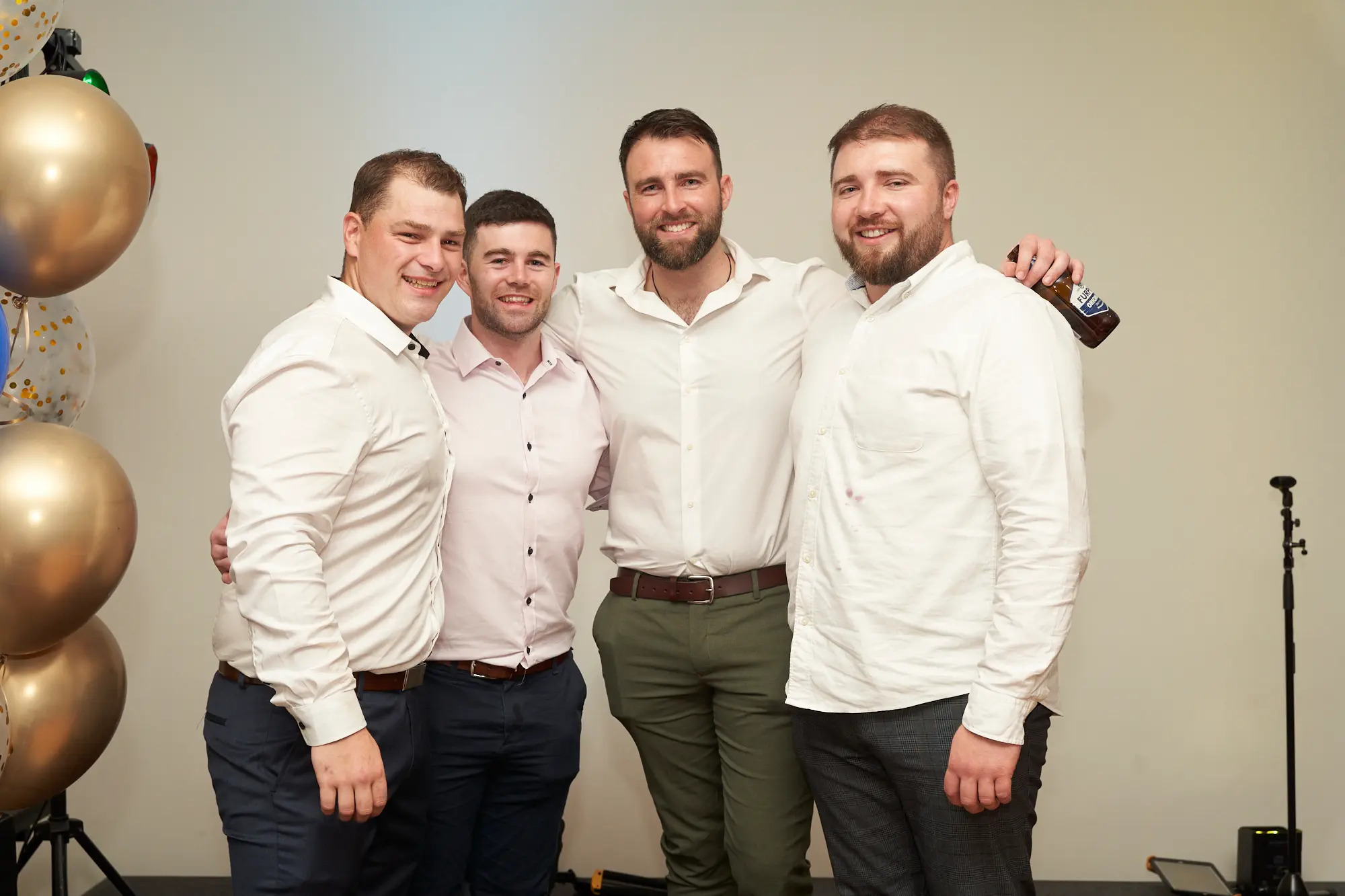 st kevins awards night pullman melbourne albert park four men group photo celebration A wide shot of four men posing together during St. Kevin’s Awards Night at Pullman Melbourne Albert Park. They stand side-by-side, smiling warmly at the camera in semi-formal evening wear—white and light pink shirts paired with dark trousers. One man holds a brown beer bottle, while another has his arm around a friend’s shoulder, showing genuine camaraderie. Behind them, gold, blue, and confetti-filled balloons decorate the beige wall, adding a festive touch. The relaxed body language and cheerful expressions reflect the fun, social, and celebratory atmosphere of the event.