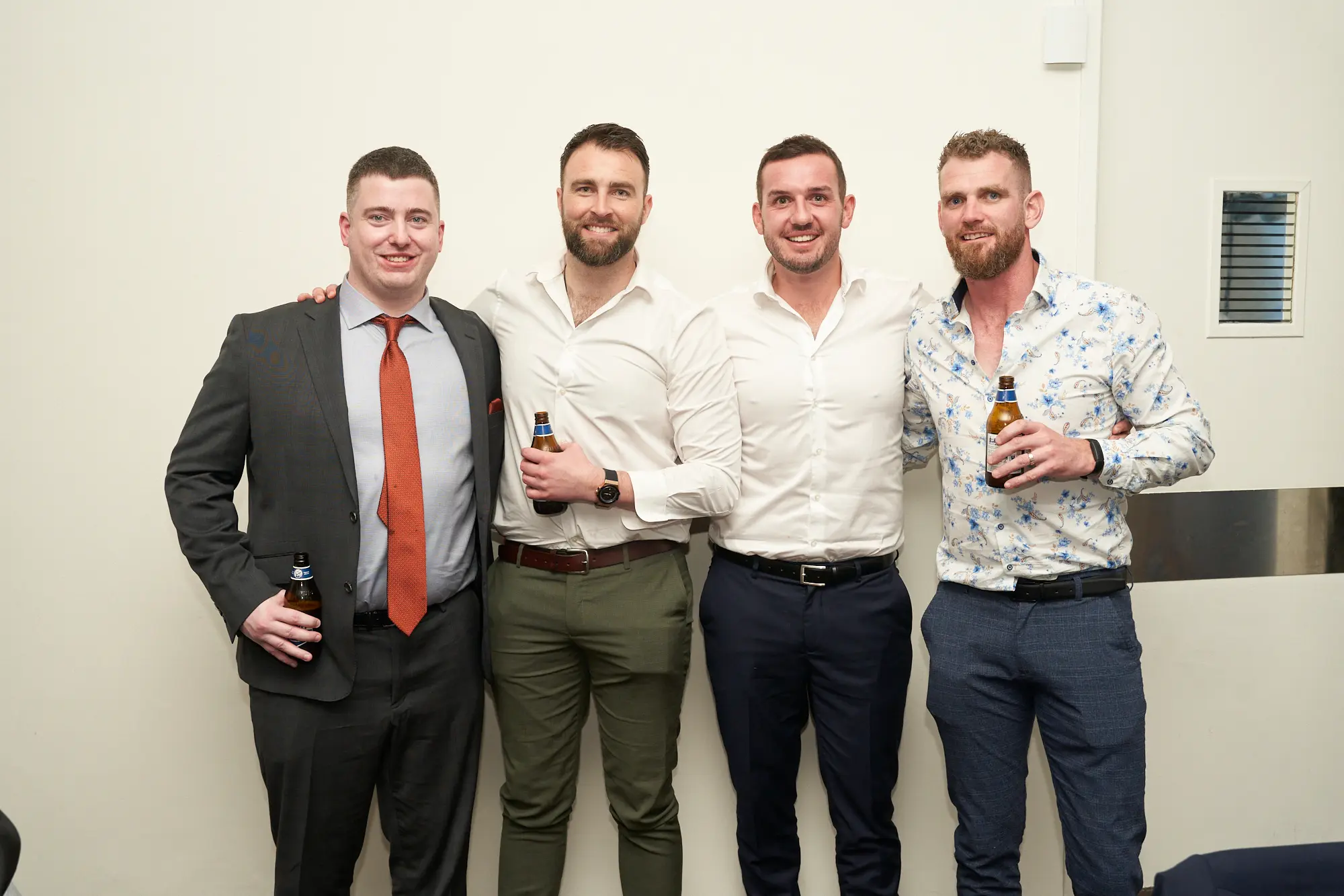 st kevins awards night pullman melbourne albert park four men group photo Four men stand side by side, smiling warmly at the camera during the St. Kevin's Awards Night celebration. From left to right: a man in a dark grey suit and rust-orange tie holds a beer, a bearded man in a white shirt and olive-green trousers holds a beer, a man in a white shirt and navy trousers has his arm around his neighbor, and a bearded man in a white floral shirt with blue-grey trousers holds a beer. The indoor setting features beige walls and bright lighting, capturing a friendly, relaxed, and festive moment among friends enjoying the event.